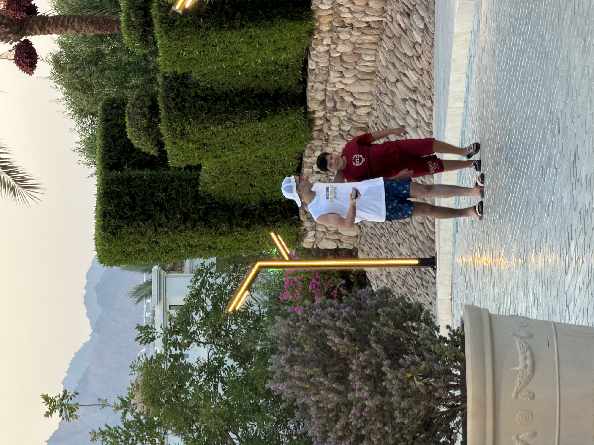 Two males are walking on a paved pathway in Sharm El Sheikh, Egypt. The male on the left is wearing a white sleeveless shirt with blue trim, blue patterned shorts, and flip-flops. He has a white baseball cap on and is holding a dark-colored mobile device in his left hand. The male on the right is wearing a red t-shirt with a graphic on the front and red shorts, and he is wearing flip-flops. They are walking side-by-side and appear to be interacting. The pathway is made of light-colored pavers and has embedded lighting that casts yellow reflections. To the left of the pathway is a large decorative planter with a shrub with purple flowers. Behind the pathway is a stone wall, followed by trimmed hedges and trees. In the background, there are buildings and mountains in the distance. The lighting suggests it is either early morning or late afternoon.