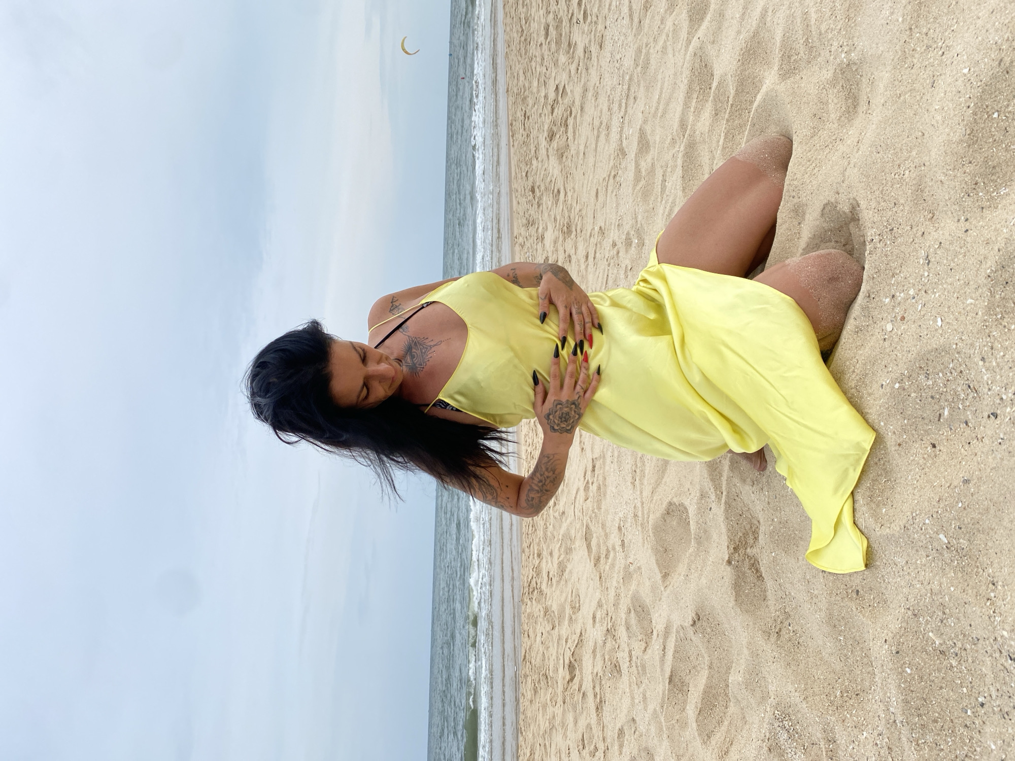 A woman with dark hair and tattoos is kneeling on a sandy beach wearing a yellow dress. The ocean is visible in the background with a kite in the sky. The location is Phu Thuy Ward, Vietnam.
