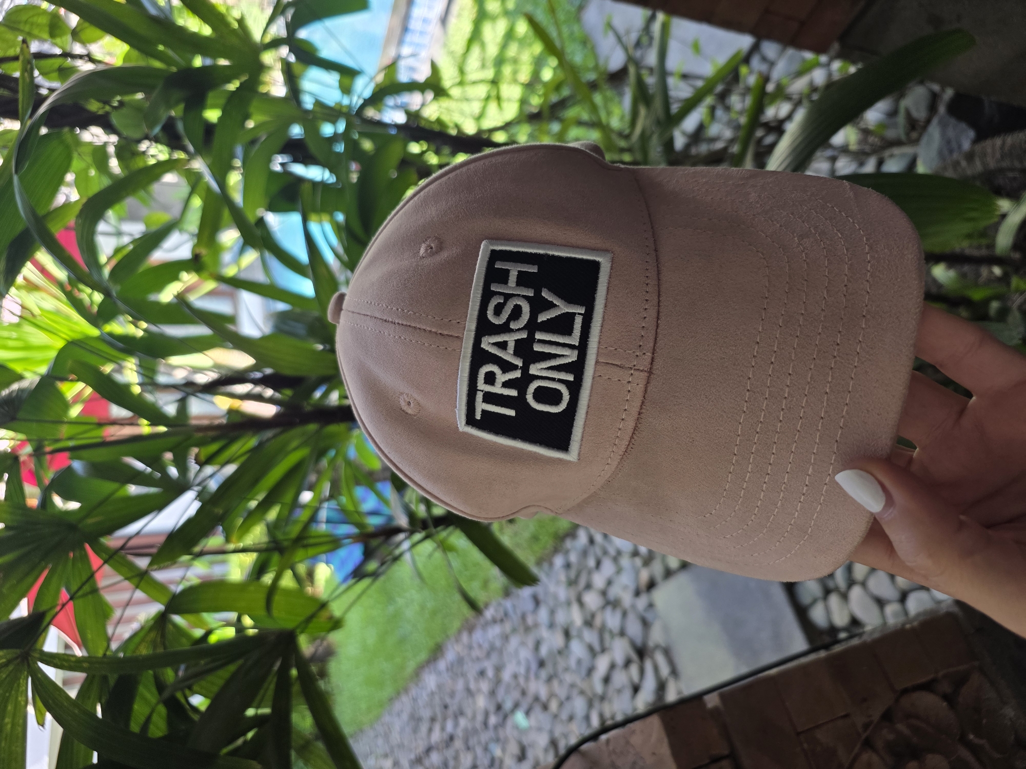 The photograph shows a tan baseball cap with a black patch that reads "TRASH ONLY" in white letters. The cap is held by a hand with a white fingernail. The background includes green leafy plants, a blue and red structure, a pebble-covered ground, and a wooden structure. Based on prior knowledge, this image was taken in North Kuta, Indonesia.
