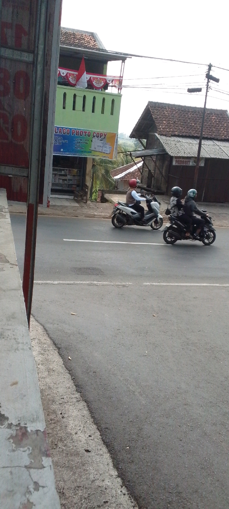 This is a vertical, medium-shot, outdoor photograph taken from a low angle of a street scene in Cilaku, Indonesia. The image captures a street with two motorcycles passing by. The motorcycle in the foreground on the right is black and carries two individuals wearing helmets. The motorcycle in the background on the left is silver and is ridden by a single individual wearing a red helmet. Both motorcycles are on the asphalt road, which has a white line painted down the center. To the left of the road is a concrete curb and sidewalk, with the side of a building visible at the far left edge of the frame. Behind the motorcycles and across the street are several buildings. One building, painted green, has a sign that reads "LAGO PHOTO COPY". Another building, with a brown tiled roof and a gray corrugated metal awning, is also visible. The sky is overcast, and palm trees are visible in the background, suggesting a tropical or subtropical climate. The overall impression is of a typical street scene in a semi-urban or rural area in Indonesia.