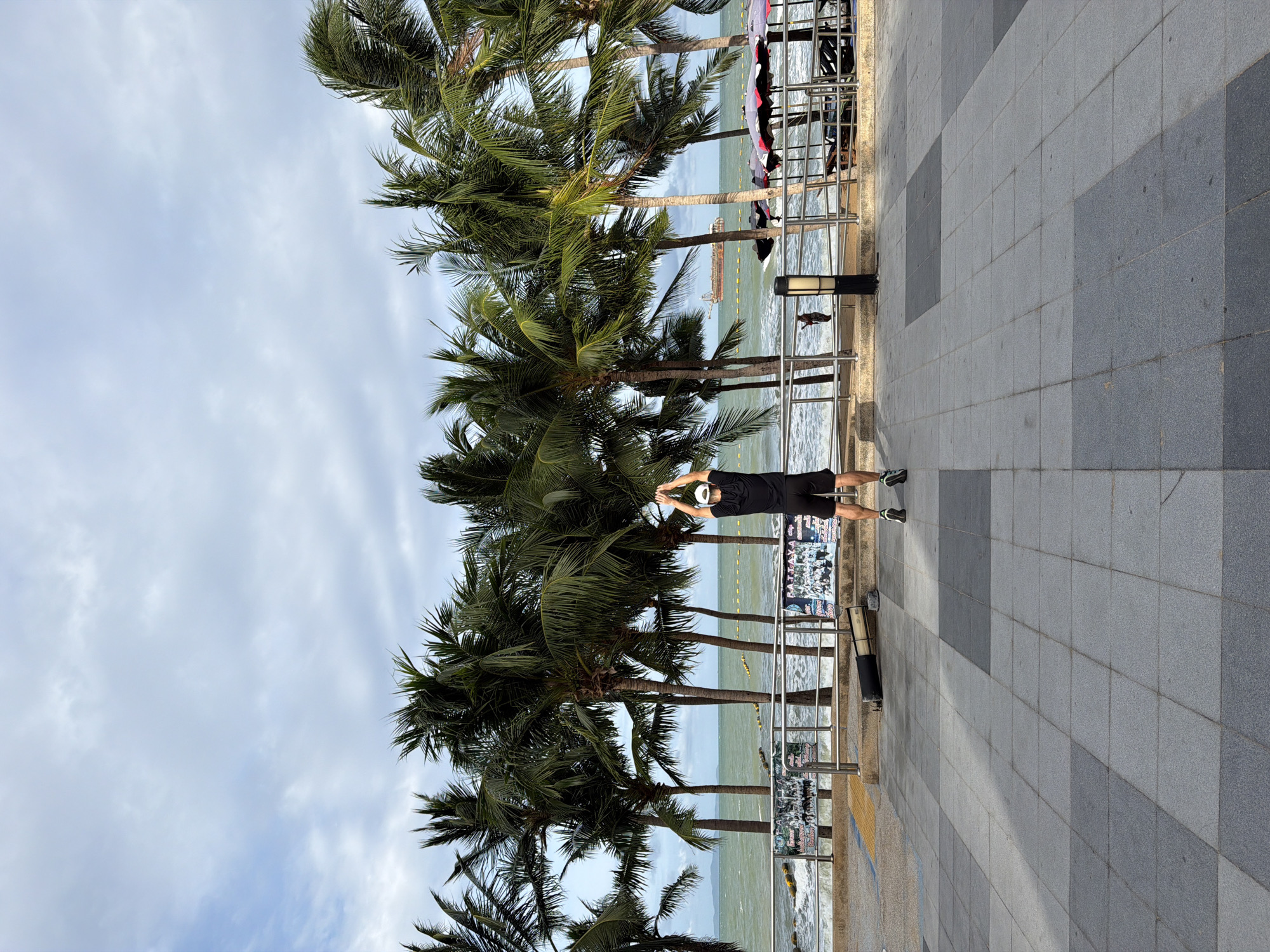 The image depicts a daytime scene on the promenade of Pattaya City, Thailand, overlooking the ocean under an overcast, light blue-grey sky. A paved pedestrian walkway composed of alternating light and dark grey square tiles occupies the foreground. In the center, facing away from the camera, a person wearing a black t-shirt, black shorts, a white cap, and athletic shoes stands with arms raised overhead, appearing to stretch or exercise. To the left of this person, a black cylindrical object, possibly a speaker or equipment case, lies horizontally on the promenade.
A silver metal railing runs horizontally across the midground, separating the promenade from the beach and ocean. Beyond the railing, the sea displays greenish-blue water with visible white-capped waves crashing towards the shore. A line of yellow buoys extends across the water in the middle distance. Several tall, slender palm trees, primarily coconut palms, form a dense line directly behind the railing, obscuring parts of the shoreline and distant elements.
Through the gaps in the palm trees and railing, a large, dark brown ship or barge is visible on the horizon towards the right side. On the far left horizon, faint landmasses or mountains are present. On the beach to the right, behind the railing, several red and white patterned beach umbrellas are set up with accompanying beach chairs. A person on a jet ski is visible in the water near these umbrellas. Further to the left, behind the railing, small figures of people are present on the beach near the water's edge. Two streetlights with dark bases and light-colored fixtures are positioned along the railing; one is partially visible on the far right, and another is more centrally located. Several Thai-language posters or advertisements are affixed to the railing supports on both the left and right sides.