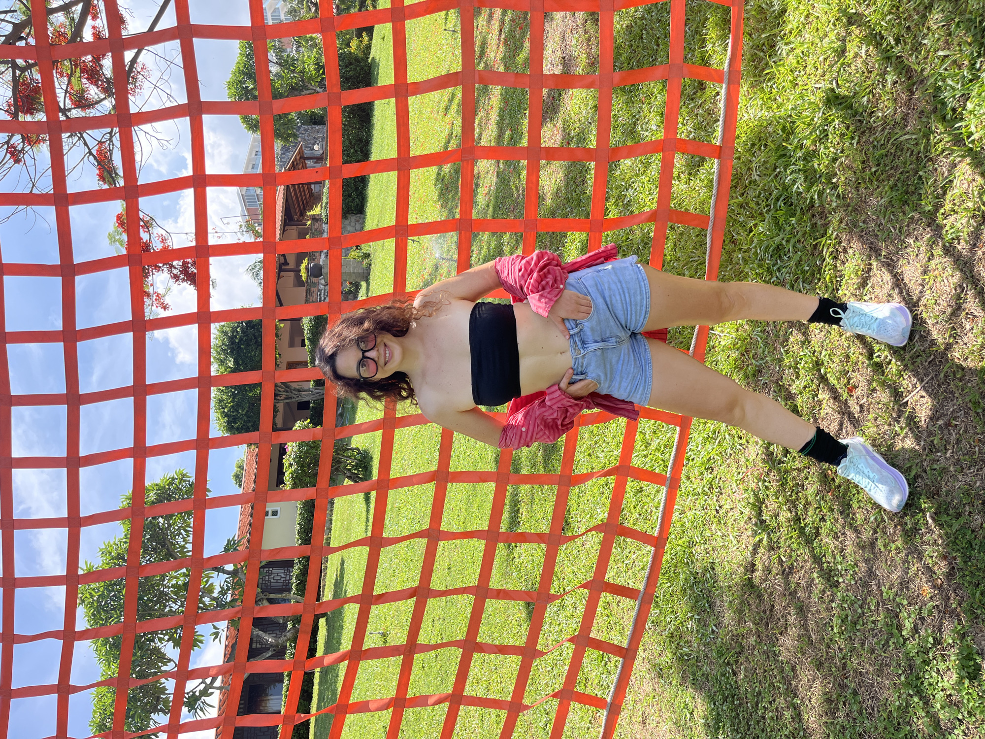 A woman with curly brown hair and glasses is posing on a large orange net. She is wearing a black tube top, denim shorts, black socks, and light blue sneakers. A pink shirt is draped over her shoulders. The net is set up outdoors, with a grassy area and trees in the background. The sky is blue with some clouds, suggesting it is daytime and fair weather. The woman is smiling at the camera, appearing happy and relaxed. The scene seems to be a recreational area, possibly a park or an outdoor attraction. Visible text is not present in the image.