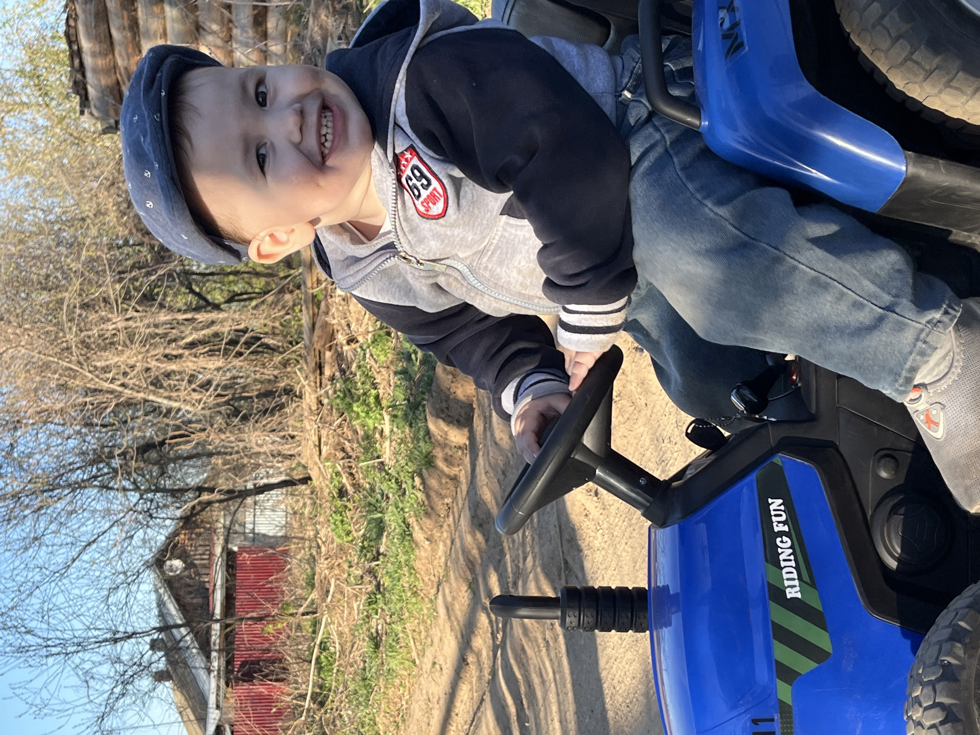 A young child, likely a toddler, is seated on a blue toy tractor in an outdoor setting. The child is wearing a blue cap, a grey hooded sweatshirt with a "69 Sport" patch, and blue jeans. The child is smiling, with the steering wheel in front of them. The tractor has "RIDING FUN" printed on the side. The background includes a dirt ground, trees, and a building with a red roof. The location is near Фоки, Russia.