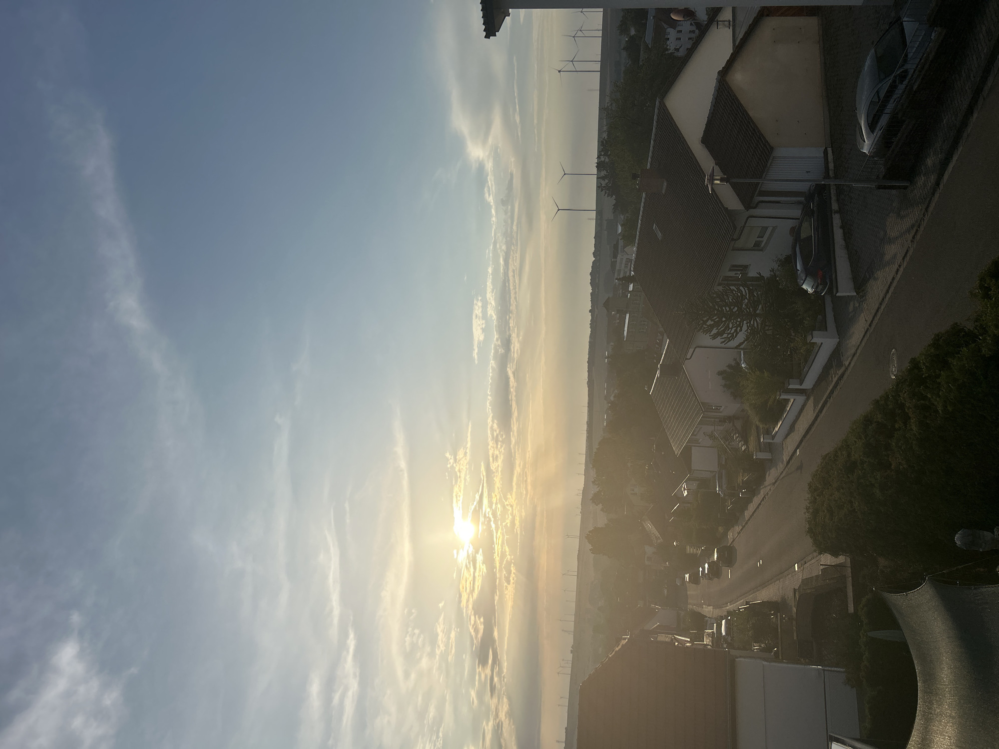 This is a high-angle, medium shot of a suburban neighborhood at sunset, with a view of wind turbines in the distance. The sun is setting on the left side of the frame, casting a warm glow over the scene and creating visible light rays. The sky is blue with scattered white clouds. In the foreground, there are rooftops of houses and a street with several cars. Further in the background, there are more houses and a line of wind turbines on a hill. The overall impression is one of a peaceful, rural or semi-rural setting during golden hour.