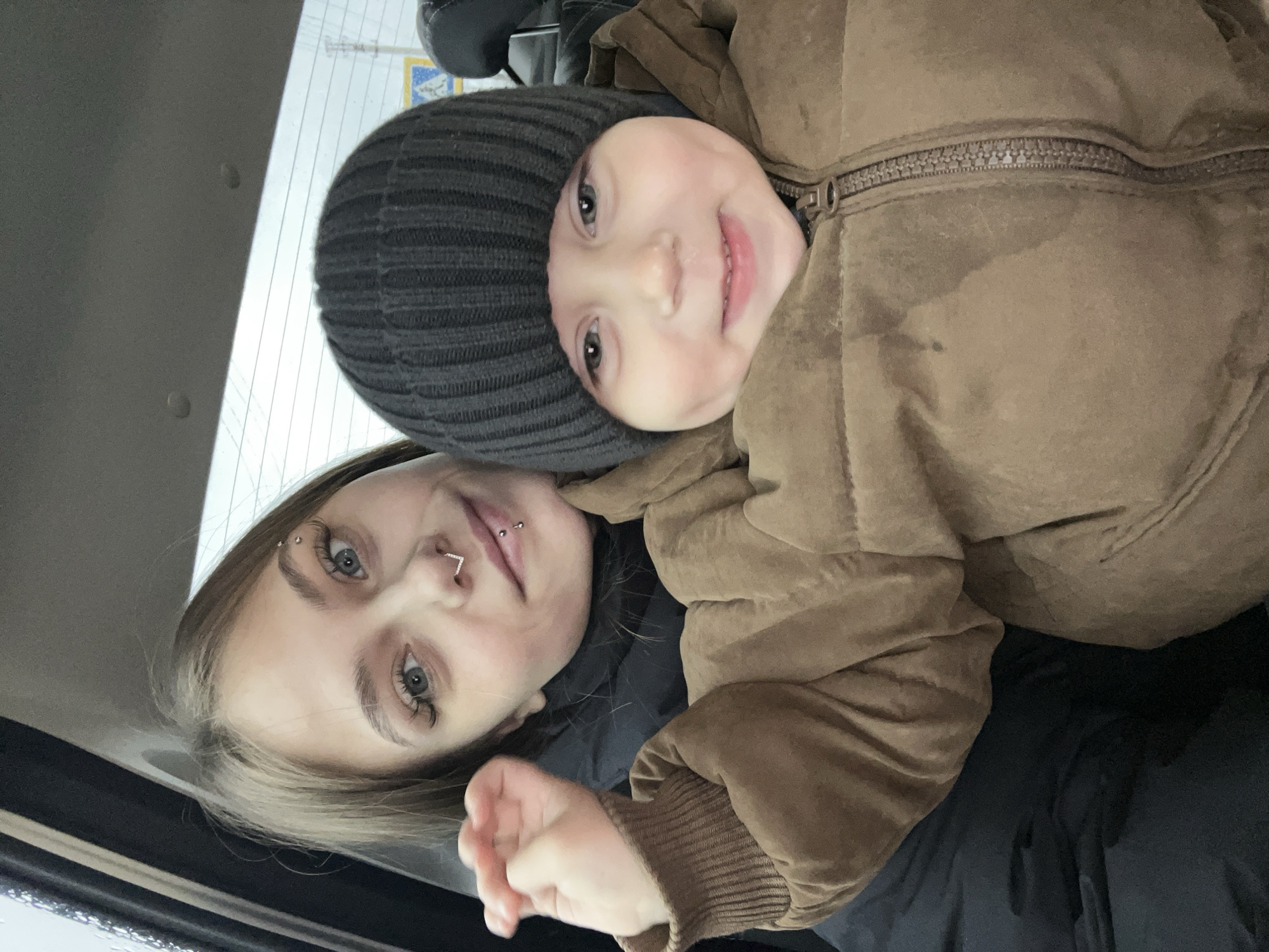 Two individuals, an adult woman and a young child, are captured in what appears to be a close-up selfie or portrait within a vehicle, possibly a car, given the visible interior elements and a rear window in the background. The image is oriented vertically, with the woman's head towards the bottom and the child's head towards the top.
The young woman, positioned closer to the camera, has light brown hair and striking blue eyes with long eyelashes. She sports several facial piercings: a horizontal barbell in her left eyebrow, a V-shaped ring in her septum, and a barbell piercing above her upper lip (philtrum). She wears a dark top, possibly a jacket, and exhibits a calm, slight smile.
To her right and slightly behind her, a young child, likely a toddler or preschooler, looks directly at the camera with a wide, joyful smile, revealing rosy cheeks. The child has light-colored eyes, possibly blue or grey, similar to the woman. They are dressed warmly in a dark grey ribbed beanie hat and a brown puffer jacket with a prominent zipper running down the front. The jacket's material appears soft and somewhat worn.
The background is softly blurred but reveals a window with faint lines, possibly defroster elements, and a subtle hint of a blue and yellow emblem or sticker. The lighting suggests it is daytime. Both individuals appear to be in good spirits, with the child's bright smile conveying clear happiness and affection. No discernible text is visible within the image itself, beyond the possible blurred emblem. The geographical context for this moment is Фоки, Russia.