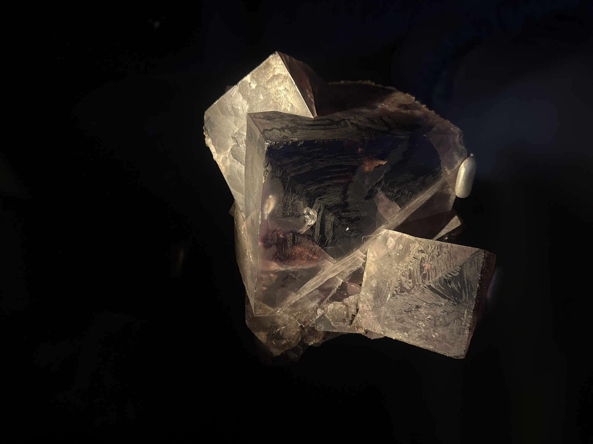 The image shows a close-up view of a cluster of amethyst crystals against a dark, indistinct background, possibly within a display case. The crystals exhibit varying shades of purple and are translucent, with light reflecting off their faceted surfaces. Some crystals are sharply defined cubes, while others are irregular clusters. The lighting is dramatic, highlighting the texture and clarity of the minerals. There is no indication of any specific location in London, United Kingdom, or any people or actions.