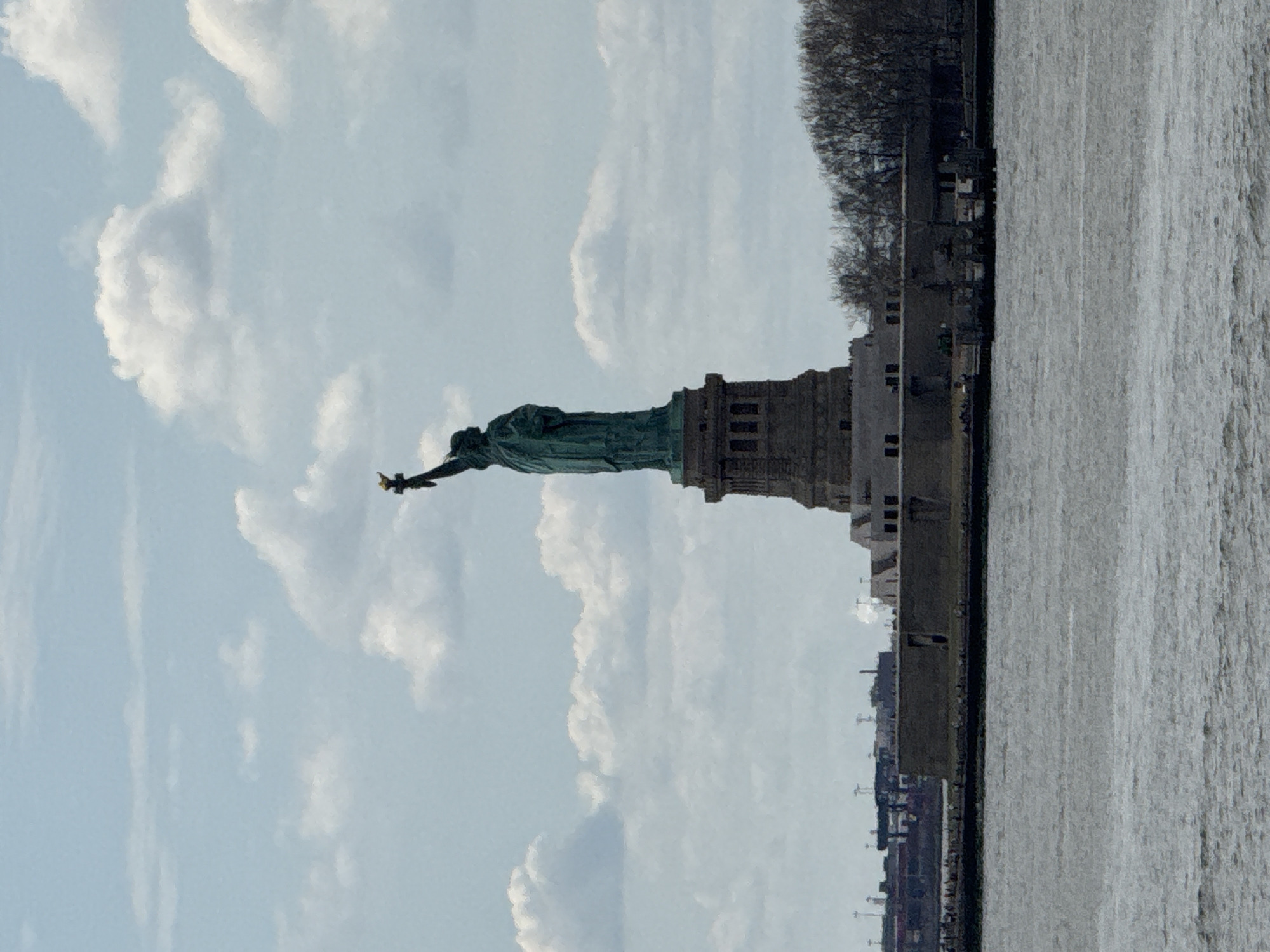 The image shows the Statue of Liberty from a distance, positioned on the left side of the frame against a cloudy sky. The statue is green and stands atop a gray pedestal structure. To the right of the statue, the image captures a shoreline or a part of an island, with a row of trees and some buildings. The water body occupies the right portion of the image. The image seems to be taken on a clear day with the sun shining. While the specific location is not evident without additional information, it suggests a view across a body of water toward Liberty Island, which is visible from Jersey City, New Jersey.