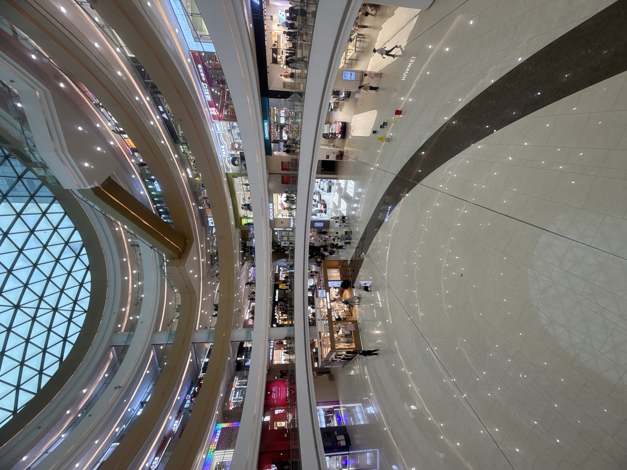 The image depicts the interior of a modern, multi-level shopping mall with a distinctive glass ceiling. The mall features several levels with shops and escalators visible. The floor is polished and reflective, with scattered lights and a dark, curved pathway. In the foreground, a person in a dark uniform walks across the floor, and further back, several individuals are scattered throughout the mall. Shops with illuminated signage are visible on multiple levels, including what appears to be a McDonald's restaurant. The architecture is contemporary, with curved balconies and integrated lighting. The scene is brightly lit, indicating it is daytime, and the overall impression is one of a bustling commercial space. The mall is located in the city of Qinhuangdao, China.