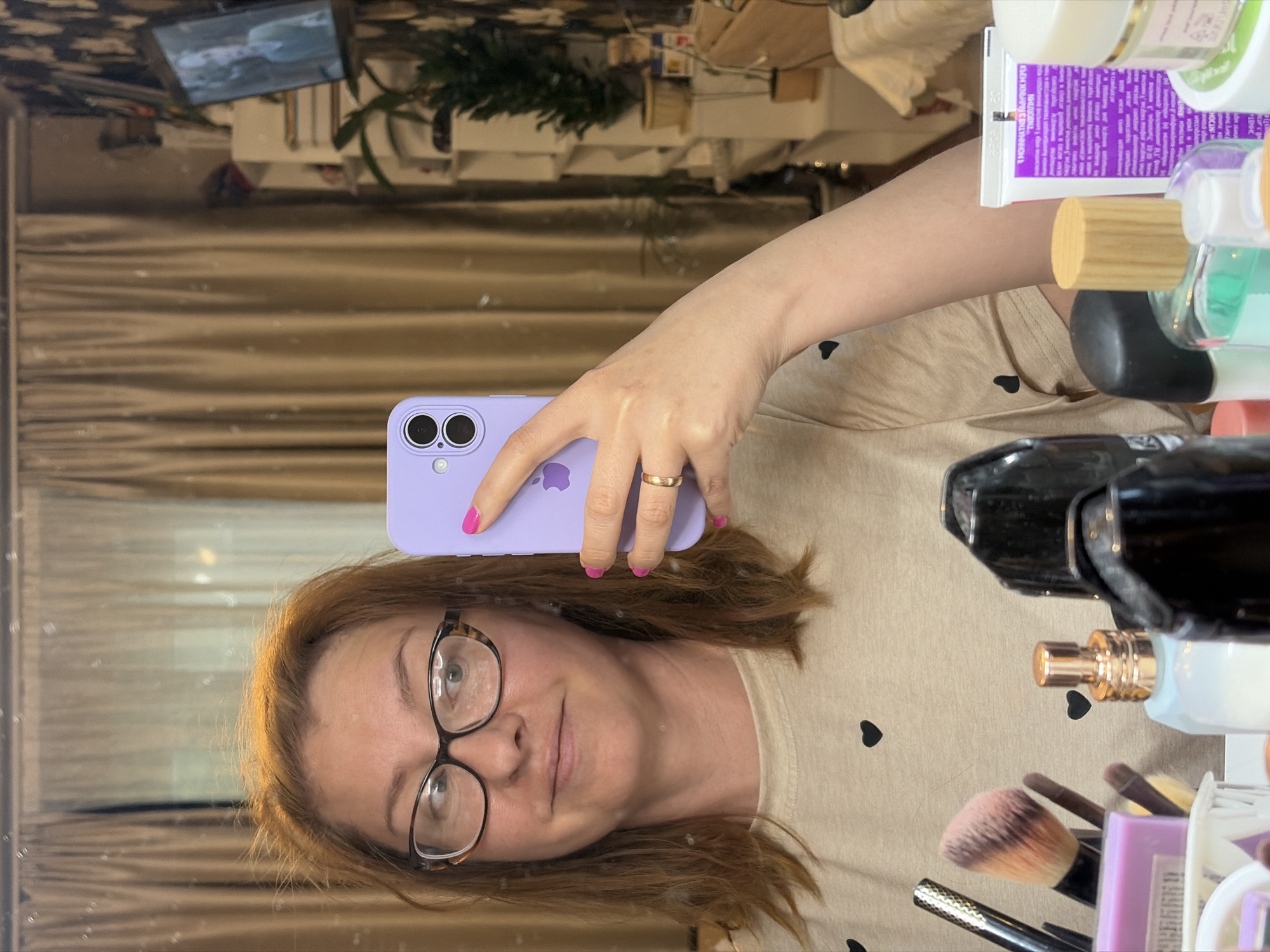 A woman with shoulder-length brown hair and glasses is taking a selfie in a mirror. She is wearing a light-colored t-shirt with small black hearts printed on it. Her left hand is holding a purple smartphone, which is pointed towards the mirror. She has bright pink nail polish on her fingernails and is wearing a ring on her left ring finger. The background is cluttered with various items, including a black hairdryer, makeup brushes, perfume bottles, and what appears to be a tube of cosmetic cream. In the reflection of the mirror, a shelf unit with some items on it, and a small television screen displaying an image, are visible. The lighting suggests it is daytime. The overall setting appears to be a dressing table or vanity area.