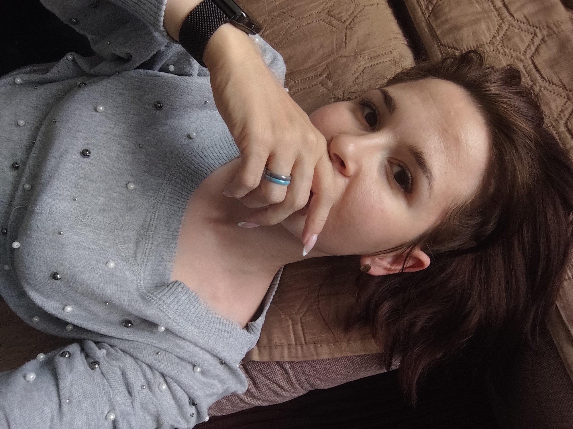 A young woman is captured in a close-up, relaxed pose, lying down on a textured brown surface, likely a couch or bed, with a quilted pattern. Her head is tilted, and she looks directly towards the viewer. She has dark brown hair, which frames her face and partially obscures her right ear, where a small earring is visible. Her facial expression is neutral or slightly contemplative.
She is wearing a light grey, long-sleeved sweater adorned with numerous small white and silver/grey spherical beads, scattered across the fabric. Her left arm is bent, and her hand is brought up towards her face, with fingers gently touching her mouth and chin area. On her left ring finger, she wears two rings: one silver/grey and one light blue. Her fingernails are manicured with white French tips. A black mesh-style watch band is visible on her left wrist, suggesting she is wearing a smartwatch. The lighting is soft and consistent with an indoor setting. There is no visible text in the image. The location context for this media file is Фоки, Russia.