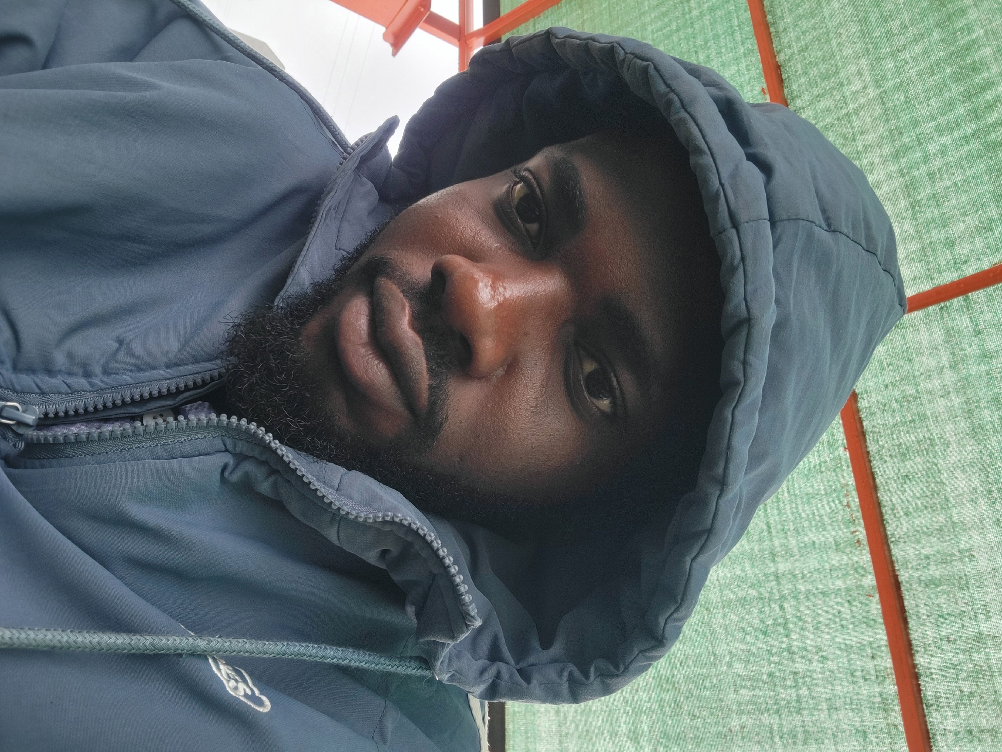 A close-up, low-angle shot captures a man wearing a dark blue hooded jacket. He has dark skin, a beard, and is looking directly at the camera. The background is a blurry green mesh with orange framing, suggesting an outdoor setting, possibly under a canopy or shade structure, in Gura Top, Nigeria. The lighting is diffused, and the man's face appears slightly shiny, indicating perspiration.