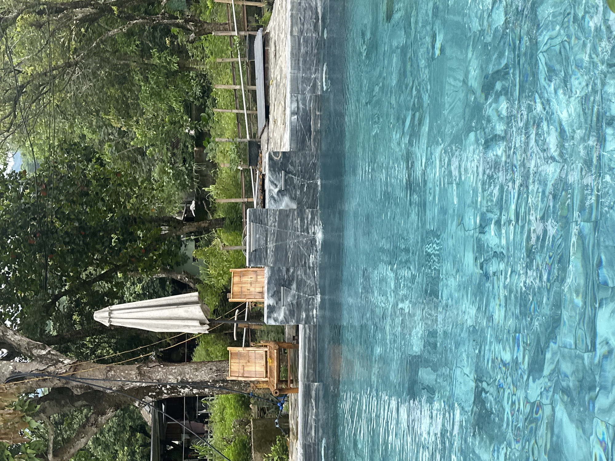 The image depicts a partially submerged swimming pool with clear, turquoise water. The pool is bordered by a stone ledge, behind which is a multi-tiered water feature made of dark, veined stone, with water cascading down. Two wooden chairs with woven seats are placed on the stone ledge, to the left of the water feature. A closed, light-colored umbrella stands behind the chairs. Beyond the pool and ledge is a lush, green environment with mature trees, dense foliage, and a bamboo fence. In the background, a rustic structure with a dark roof is partially visible among the trees. The overall scene suggests an outdoor recreational area in a natural setting. The location is identified as Xóm Khán, Vietnam.