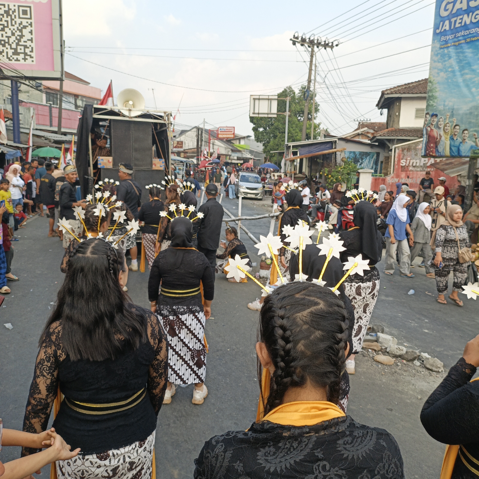 The image depicts a street scene in Randudongkal, Indonesia, during what appears to be a cultural event or parade. In the foreground, several individuals are shown from behind, dressed in traditional attire. The women wear black lace tops with intricate patterns, paired with batik skirts and golden sashes. They also have elaborate hairstyles and headpieces adorned with white, star-shaped ornaments.
The middle ground reveals more participants in similar traditional clothing, some of whom are smiling and interacting. A sound system mounted on a vehicle is visible, suggesting the presence of music or announcements. People are gathered along the sides of the street, observing the procession. The street itself is paved and shows signs of wear, with some debris scattered on the ground. Buildings line the street, with shops and banners indicating a commercial area. Overhead, power lines crisscross the sky, a common sight in urban Indonesian settings. The overall atmosphere seems festive and communal, with a sense of local pride and celebration.