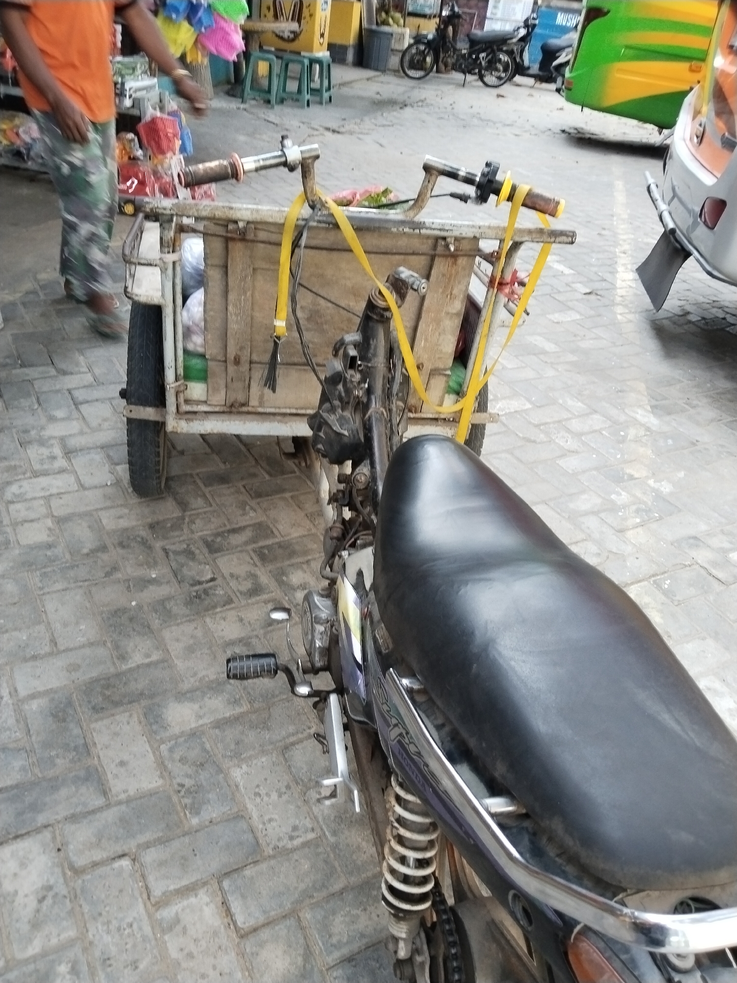 The image shows a street scene in the city of Prigi, Indonesia. A person wearing an orange t-shirt and camouflage pants is visible on the left side of the frame, walking away from the camera. In the center of the image, a two-wheeled cart is attached to a motorcycle. The cart is made of wood and appears to be carrying some items. The motorcycle has a black seat and is parked on a brick-paved street. In the background, there are some green and yellow vehicles, and other street elements that suggest a market or a busy street. The overall scene depicts a typical street environment in a city in Indonesia.