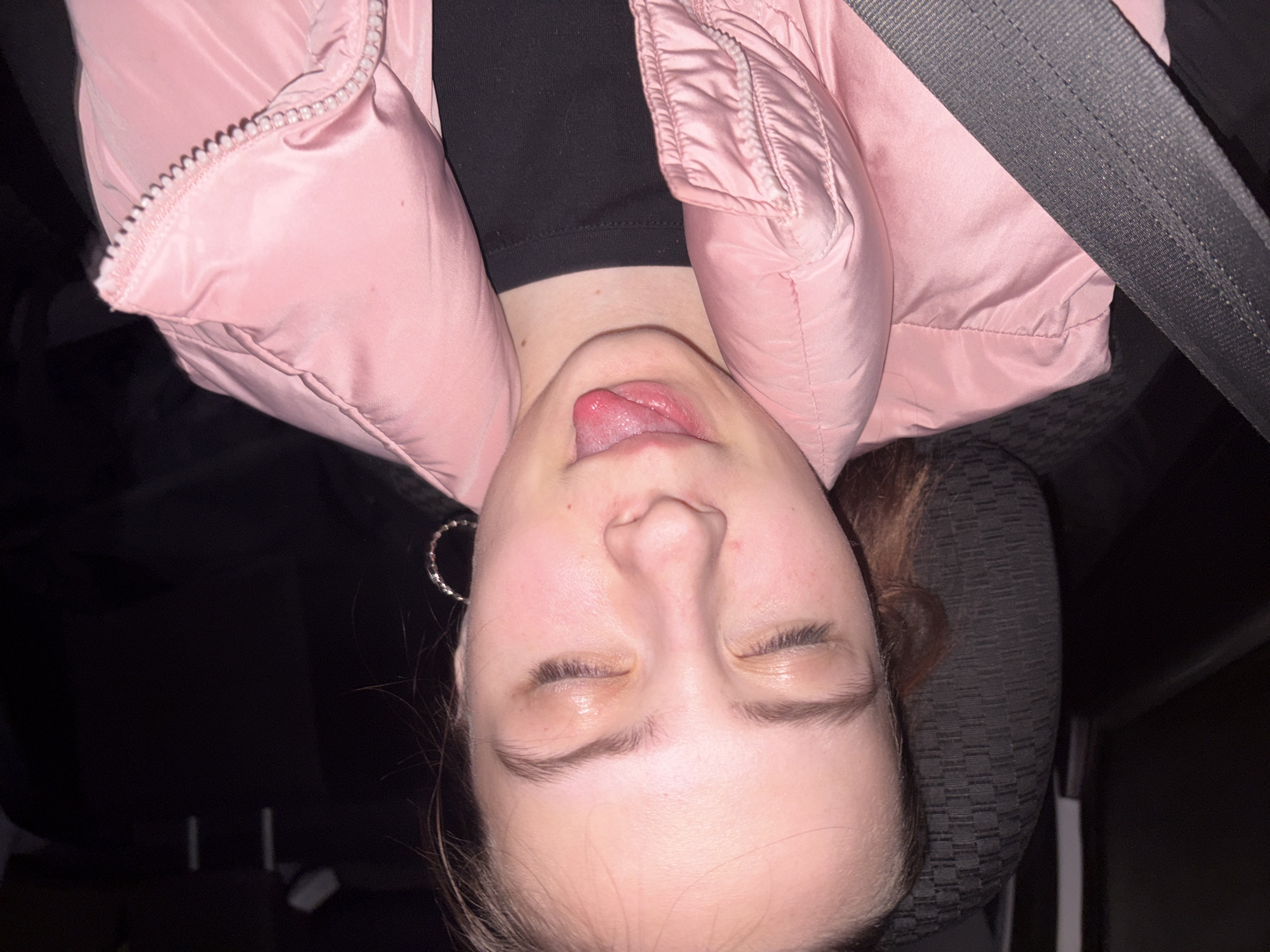 The image features a close-up portrait of a young person, likely female, situated inside a vehicle. She is wearing a light pink puffy jacket with a visible zipper, layered over a black top. Her head is tilted back, eyes closed, and tongue playfully sticking out, suggesting a lighthearted or silly pose. A large, silvery hoop earring adorns her left ear. A dark gray seatbelt runs diagonally across her upper body, over the pink jacket, confirming her presence in a car or similar transport. To the right of her head, a dark-patterned car seat headrest is visible. The surrounding environment is dark and out of focus, indicating an interior setting, possibly at night or during low light conditions. The scene captures a candid, playful moment taking place in Votkinsk, Russia. No text is visible in the image.