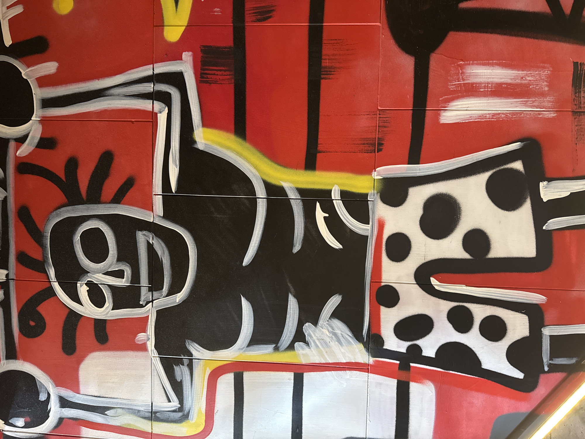 The image depicts a section of graffiti art on a tiled wall in Moscow, Russia. The dominant colors are red, black, white, and yellow. The artwork features abstract figures and shapes, with some elements resembling human-like forms. One figure on the left has a large, round head with the characters "8D" inside. The figure on the right has a white body with black dots. The overall style is reminiscent of Keith Haring's work. The tiles are visible in the background.