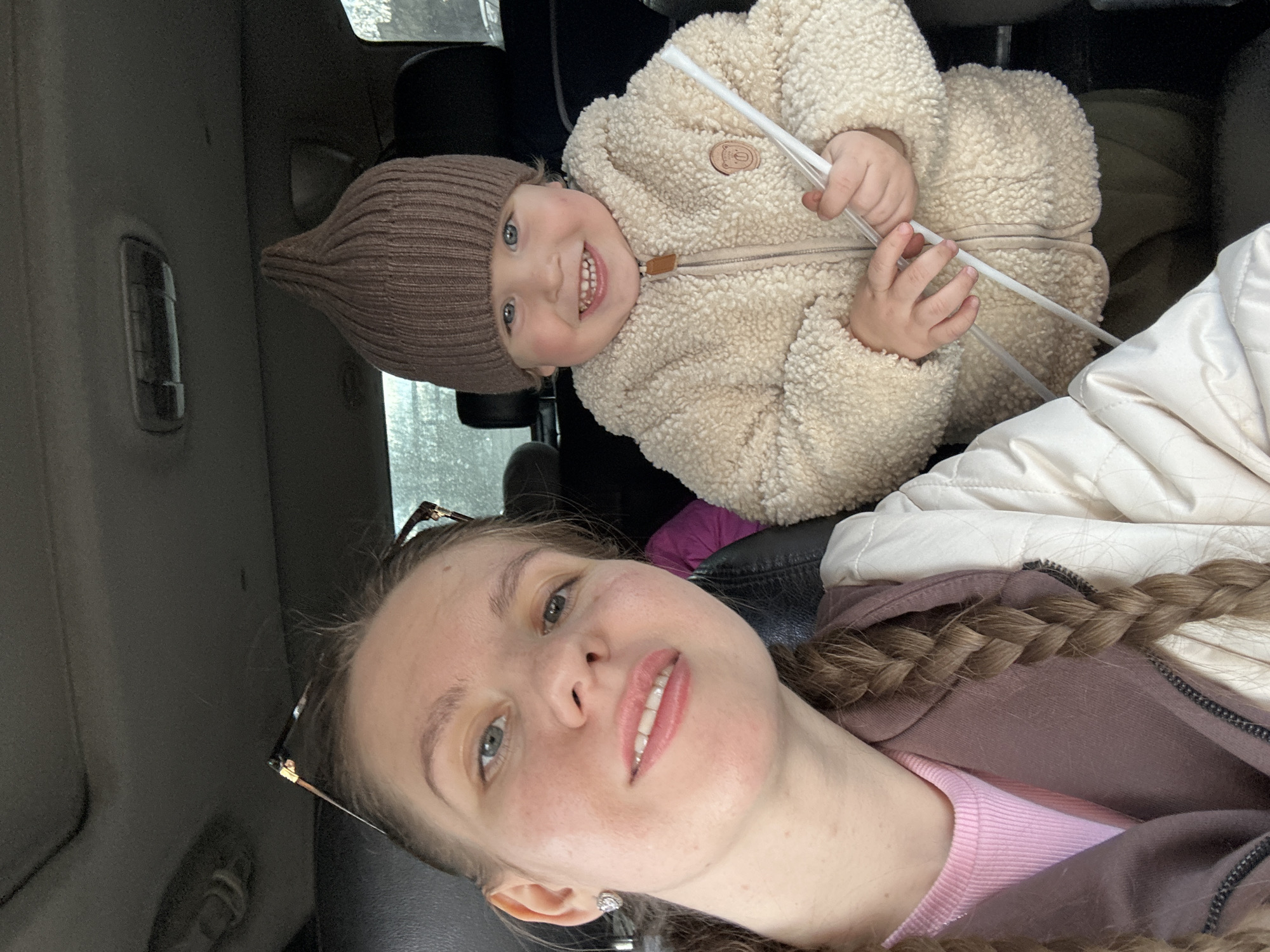 The image depicts a selfie taken inside a car. A young woman with light skin and a braid is in the foreground. She is wearing a pink top and a brown hoodie. A young child, possibly a toddler, wearing a brown knit hat and a cream-colored fleece jacket, is visible in the background. The child is holding two white sticks. The setting appears to be the interior of a car. The location is Klin, Russia.