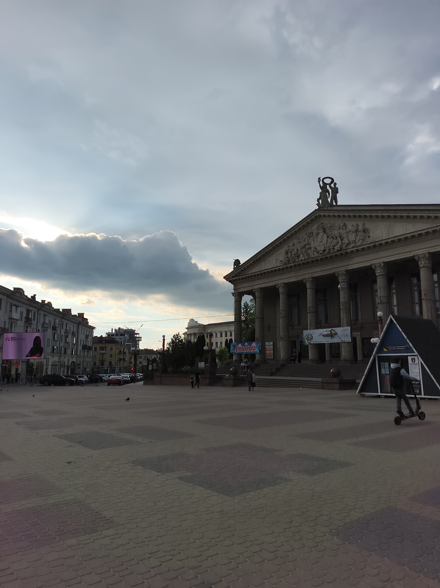The image depicts the Theater Square in Ternopil, Ukraine. The Ternopil Academic Regional Ukrainian Drama Theater named after Taras Shevchenko stands prominently on the right. The square itself is paved with grey and brown brick and features a small, triangular kiosk. Several people are visible in the image: One person is riding a scooter, and others are walking through the square. Cars line the street beyond the square to the left, along with buildings and advertising displays. The sky is overcast with a bright patch of light visible above the left side of the frame.