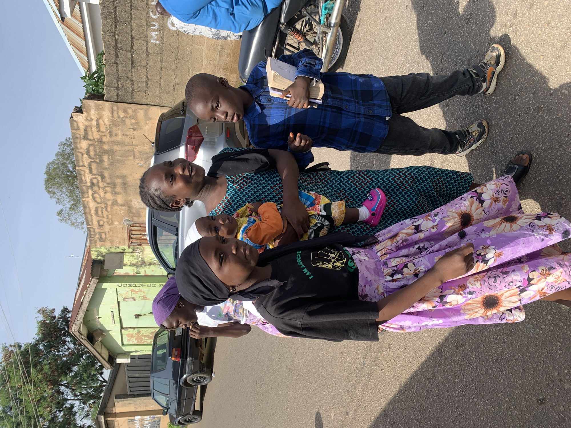 In Jos, Nigeria, a group of children stands on a paved street. The child on the far left wears a black t-shirt with a green graphic and a purple floral skirt. Next to them, a younger child in an orange and white outfit is held by a taller child wearing a teal, patterned dress. To the right, a boy in a blue plaid shirt and dark pants holds a book. Behind them, a black car is parked on the left side of the road, and a motorcycle is visible on the right. Buildings with weathered walls and corrugated metal roofs form the backdrop, with trees and a clear sky above.
