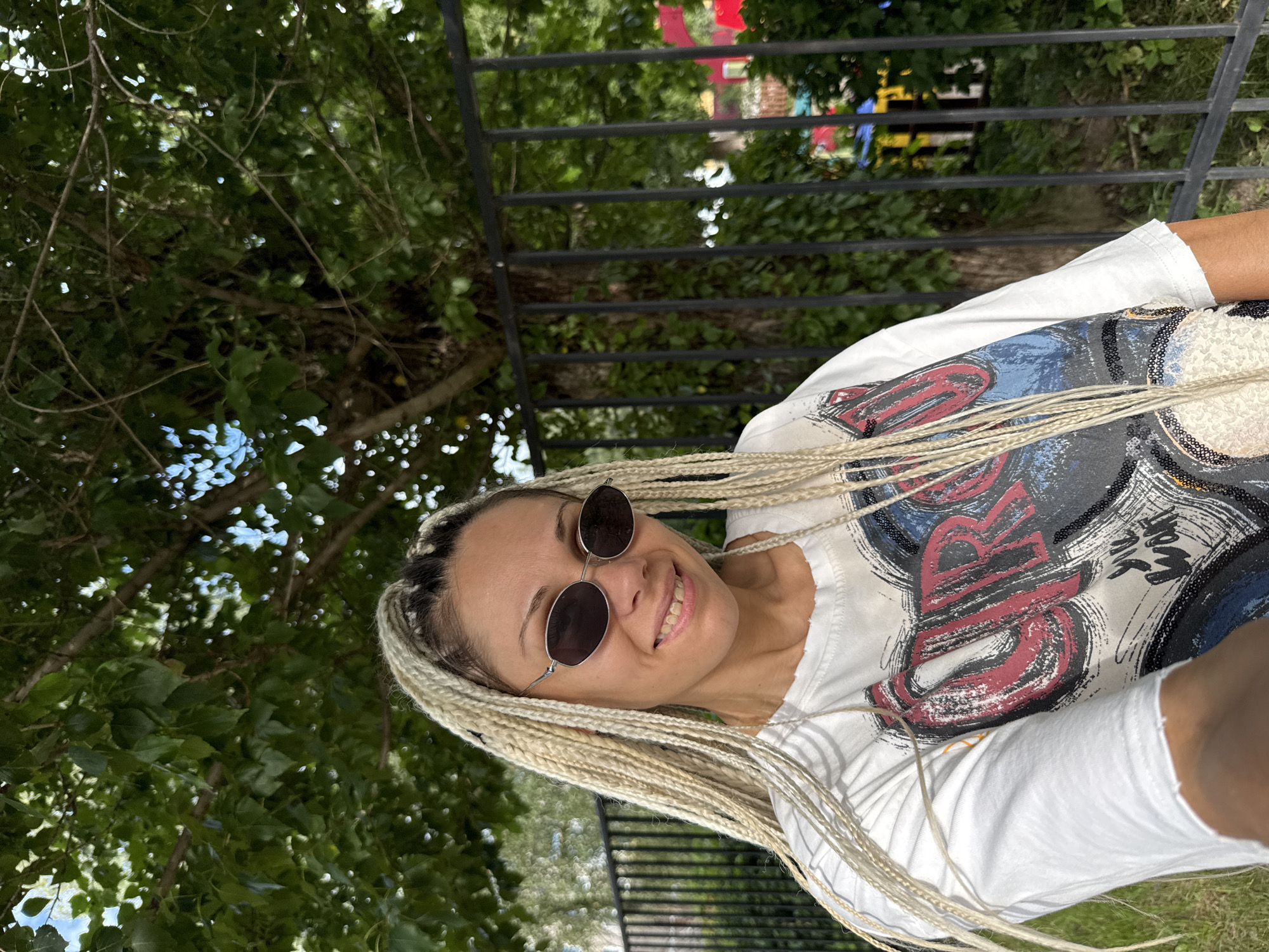 A medium shot captures a woman with blonde braided hair and sunglasses smiling at the camera, standing outdoors. She is wearing a white t-shirt with a graphic print. A black metal fence is behind her, with trees and foliage in the background. To the right of the fence, colorful structures, possibly playground equipment, are visible. The scene appears to be in a park or outdoor recreational area within the city of Rusanovo, Russia.