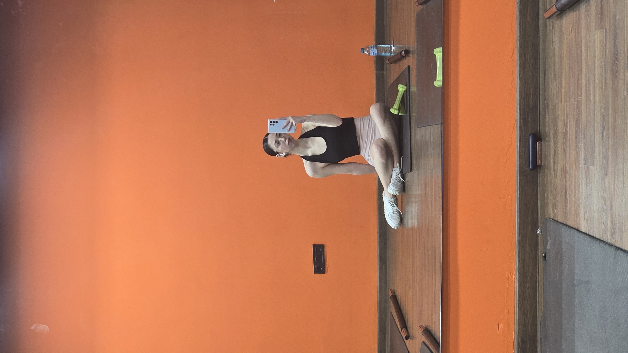 A mirror selfie in a gym setting. The image captures a woman in workout attire—a black tank top and light-colored shorts—seated on a yoga mat, holding a light blue smartphone up to take a picture of herself. Beside her are small green dumbbells and a water bottle. The background features a bright orange wall, contrasted by the wooden-paneled floor and a second yoga mat with weights. The reflection suggests this scene may be in a fitness studio or gym. Without more details, I cannot determine the exact location in Stara Zagora, Bulgaria.