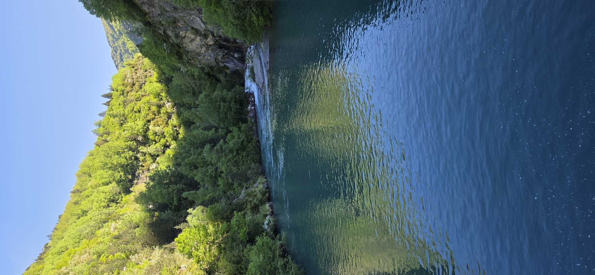 The image shows a body of water bordered by a lush, green hillside in Benasque, Spain. A small waterfall cascades down a rocky cliff face into the water. The water's surface reflects the surrounding greenery and sky, creating a rippled texture. The hillside is densely covered with trees and vegetation. The overall scene is a natural landscape with a blend of water, rocks, and greenery.