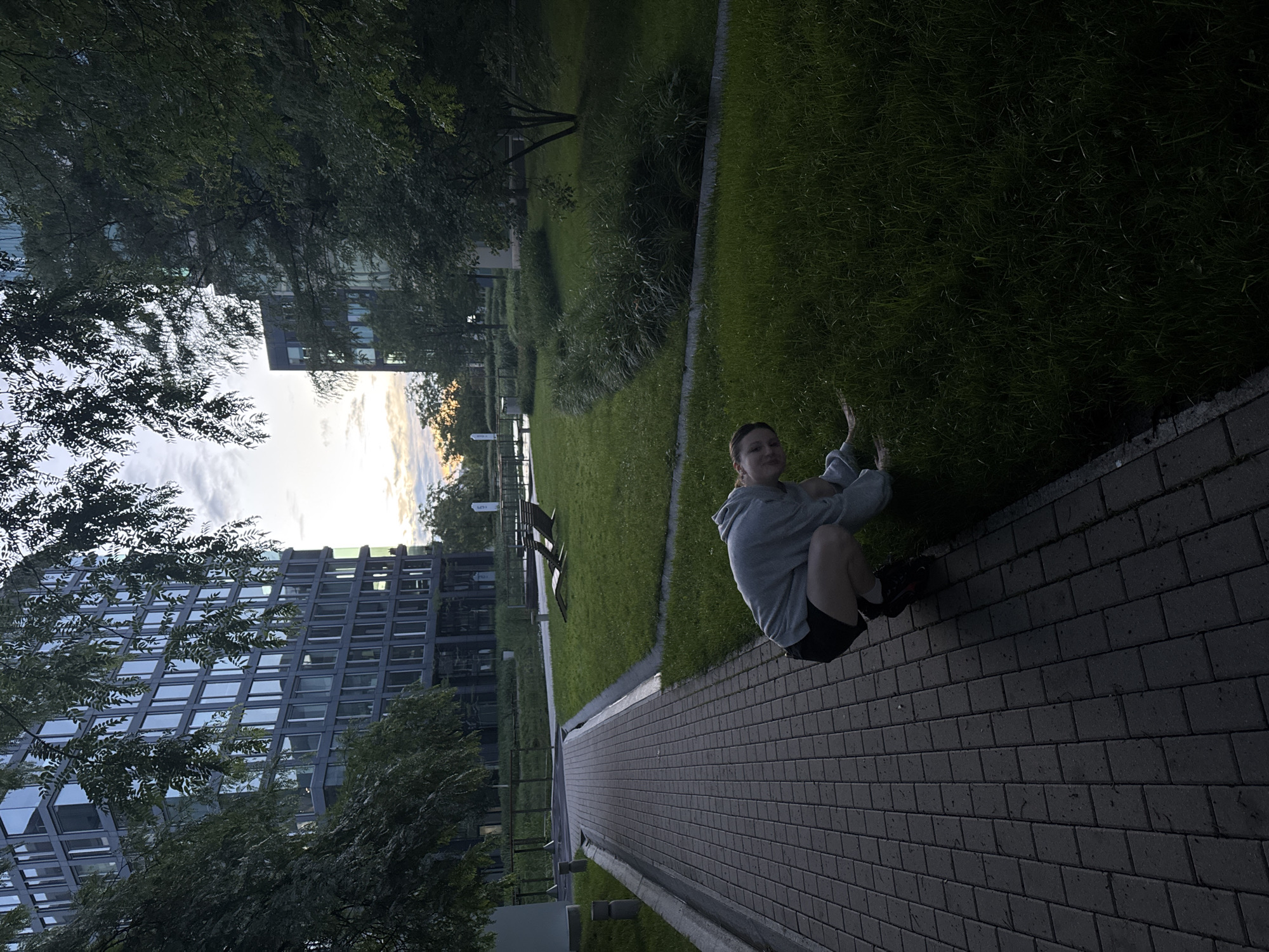 A young person is crouching on the grass next to a paved walkway. The person is wearing a gray hooded sweatshirt and black shorts. The walkway is made of dark gray pavers arranged in a basket-weave pattern. Behind the person, the grass slopes gently upwards towards several modern, multi-story buildings with large windows. Trees and bushes are visible in the foreground and between the buildings. In the background, there are a few lounge chairs on the grass. The sky is partly cloudy, with the setting sun casting a warm glow. The scene appears to be a park or courtyard area within an urban environment.