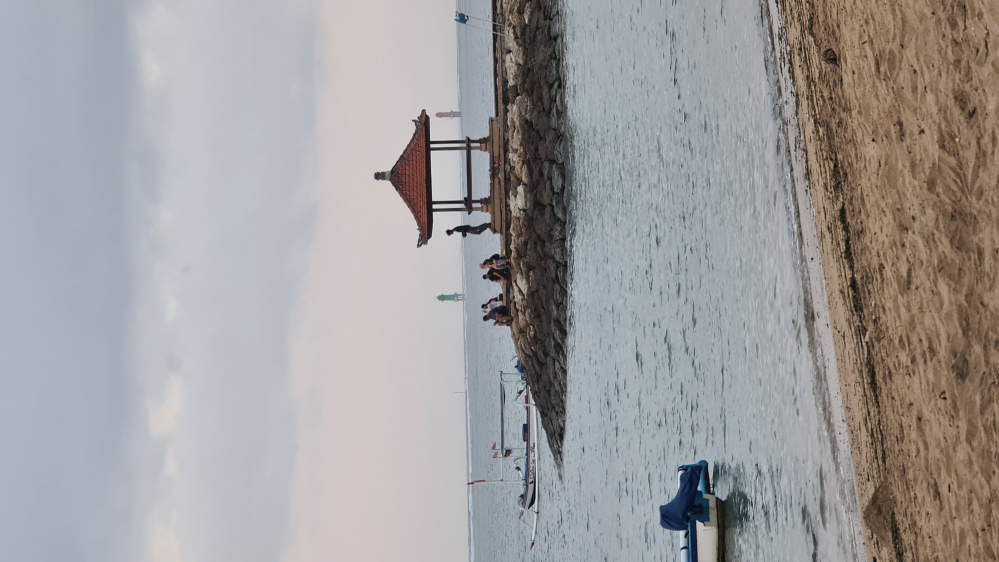 The image depicts a serene coastal scene, likely at Sanur Beach in Denpasar, Indonesia. The foreground features a sandy beach with gentle waves lapping at the shore. In the middle ground, the calm sea is dotted with several traditional Balinese fishing boats, their white hulls and outriggers contrasting with the water.
Further out, a stone jetty juts into the sea, topped by a traditional Balinese pavilion with a red tiled roof. Several individuals are seated on the jetty, some appearing to be fishing or simply relaxing. One person stands near the pavilion, possibly engaged in fishing as well. In the background, two lighthouses, one green and one red, stand as navigational aids in the hazy distance. The sky is overcast, casting a soft, diffused light over the entire scene. The overall atmosphere is peaceful and tranquil, characteristic of a quiet morning or late afternoon at a popular beach destination.