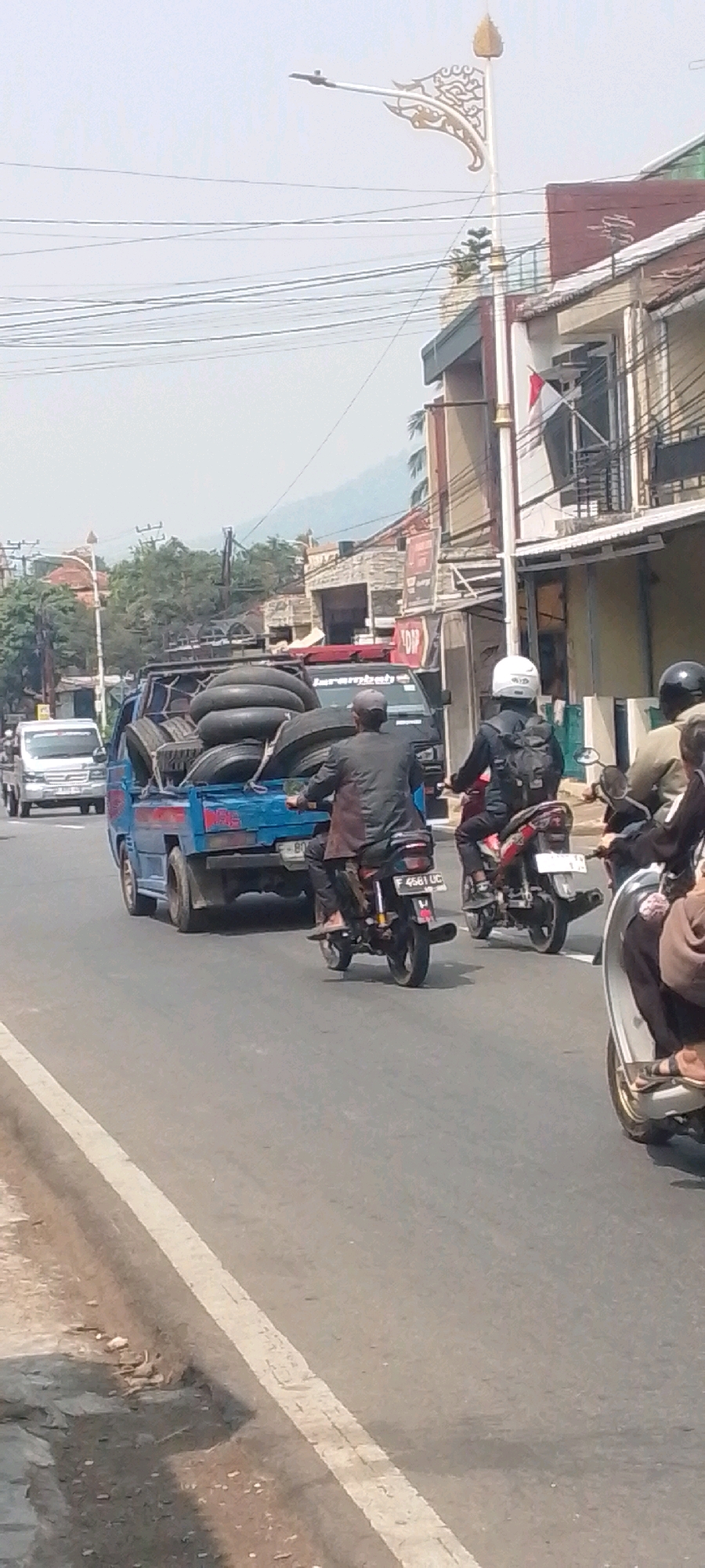A blue pickup truck, loaded with several large tires, drives down a paved road in Sukasari, Indonesia. The truck is in motion, as are several motorcycles. One motorcycle, with a rider wearing a helmet and backpack, is to the right of the truck and appears to be following it. Another motorcycle, with a rider wearing a hat, is behind the truck. The road is bordered by a white line. Buildings with balconies and awnings are visible on the right side of the road, and trees and hills are in the background. The sky is hazy.