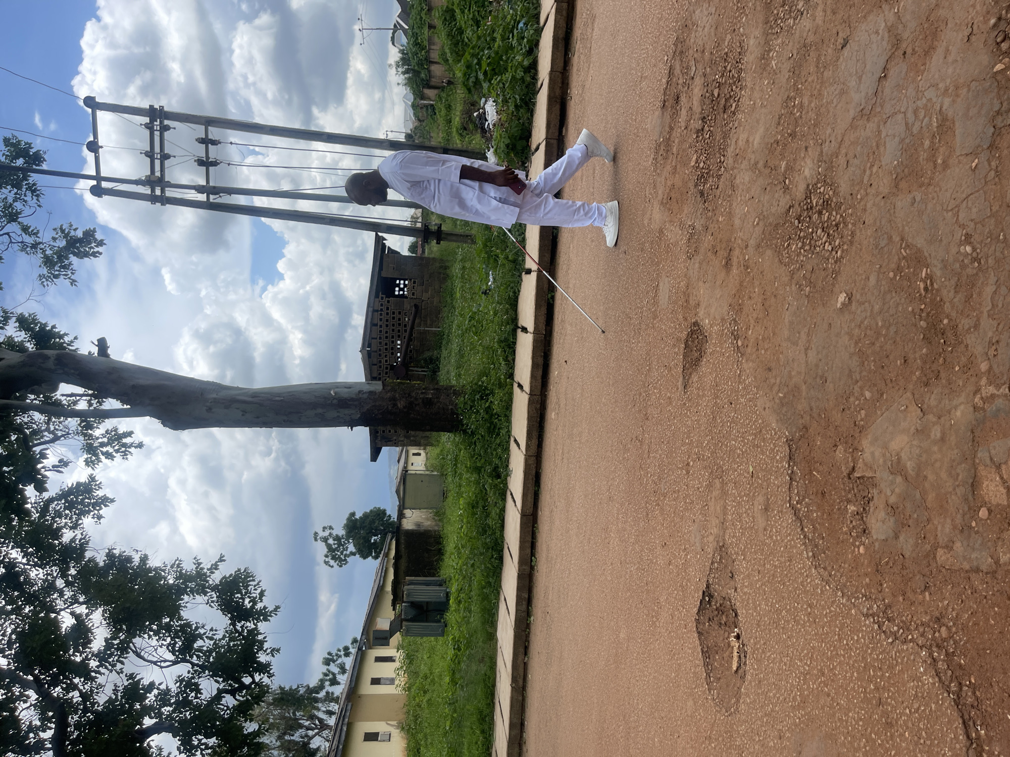 A visually impaired man wearing white traditional attire is walking on a dirt road in Rafin Par, Nigeria, using a white cane to navigate. The road is in poor condition, with several potholes visible in the foreground. In the background, there are some residential buildings, a transformer, and a large tree. A utility pole with electrical wires stands prominently on the right side of the image. The sky is cloudy.