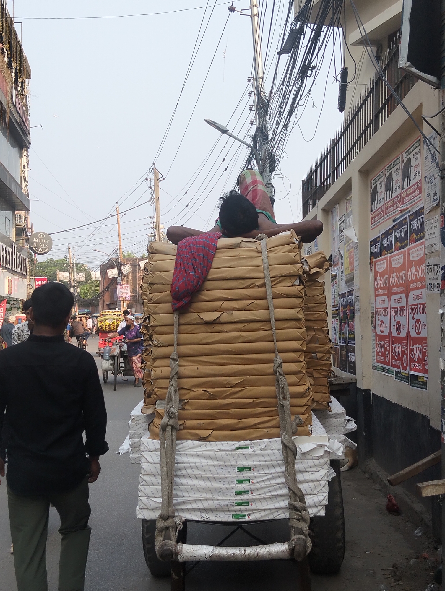 A medium shot shows a street scene in Dhaka, Bangladesh. A man is lying on top of a large load of bound paper on a cart, which appears to be pulled by a vehicle out of frame. The man is wearing a red and green patterned cloth covering his head and shoulders. The cart is stacked with bundles of brown-wrapped paper on top of white-wrapped paper. Two thick, rope-like straps are tied around the stack.
To the left of the cart, a man in a black shirt and olive-green pants is walking away from the camera. In the background, people are walking and riding bicycles and rickshaws along the street. Buildings line the street, some with signs in English and Bengali. The sky is overcast.
To the right of the cart, a wall is covered with posters in Bengali. Wires are strung overhead, creating a dense web against the sky.
