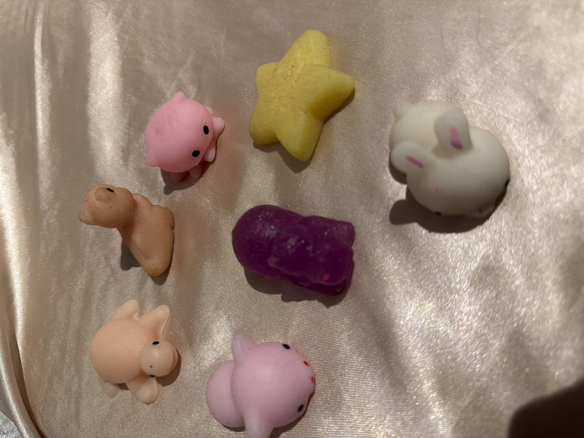 The image shows seven squishy toys on a light beige, slightly shiny fabric. There is a pink octopus, a yellow star, a white rabbit, a brown bear, a purple amorphous shape, a light peach pig, and a pink pig. The location is not discernible from the image.