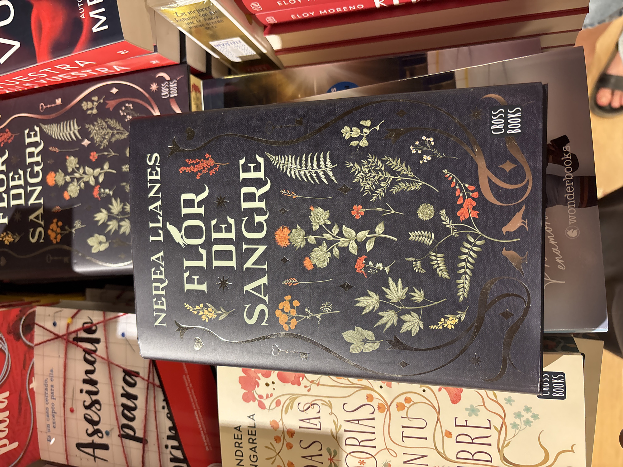 The image shows a close-up view of a stack of books in a bookstore. The main subject is a dark blue book with gold lettering and floral illustrations. The title on this book appears to be "FLOR DE SANGRE" by NEREA LLANES. A sticker on the book reads "CROSS BOOKS". To the left of this book is a cream-colored book with similar ornate illustrations and text. Further to the left, partially visible, is a red book with "ASESINATO PARA" written on it. In the background, other book spines are visible, including a red one with the name "ME" and another with "NUESTRA".
The image is a still photograph taken from a slightly elevated angle, looking down at the books. The lighting suggests an indoor environment, likely a retail setting. There are no visible people in the frame. The overall impression is that of a display of books for sale. The question about the location in the city of Orihuela, Spain, cannot be answered from the provided image as there are no identifying landmarks or text that would indicate a specific city or region.