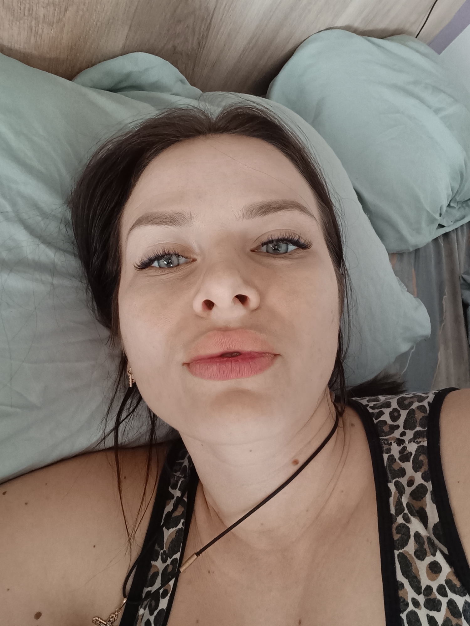 A woman lies on a bed with light blue bedding. She has dark hair and blue eyes, and she's wearing a leopard-print tank top and a thin black necklace. The background includes a light-colored wooden headboard. There is no verifiable evidence of Rusalevka, Russia.