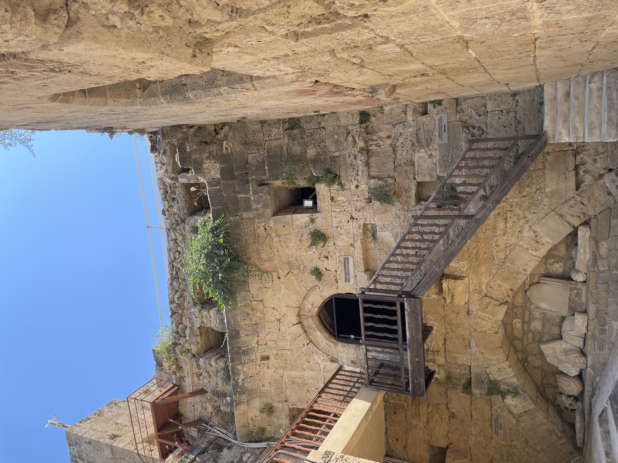 The image shows a view looking up into the courtyards of Kyrenia Castle, located in Kyrenia, Northern Cyprus. The castle is constructed of rough-hewn stone blocks, with rough masonry walls and archways. A wooden staircase with dark brown railings ascends to an arched doorway with a dark interior. Above the doorway, a small, square opening is visible in the stone wall. To the left, a lighter colored balcony with similar wooden railings extends from a tan colored structure. Above this, a section of stone wall is topped with a rusted metal platform and railing, and a cross is visible on the highest point of the structure. Overgrown green vegetation sprouts from cracks in the stone walls. In the lower section of the image, beneath an archway, several ancient-looking stone artifacts are displayed. The sky above is a clear, bright blue.
