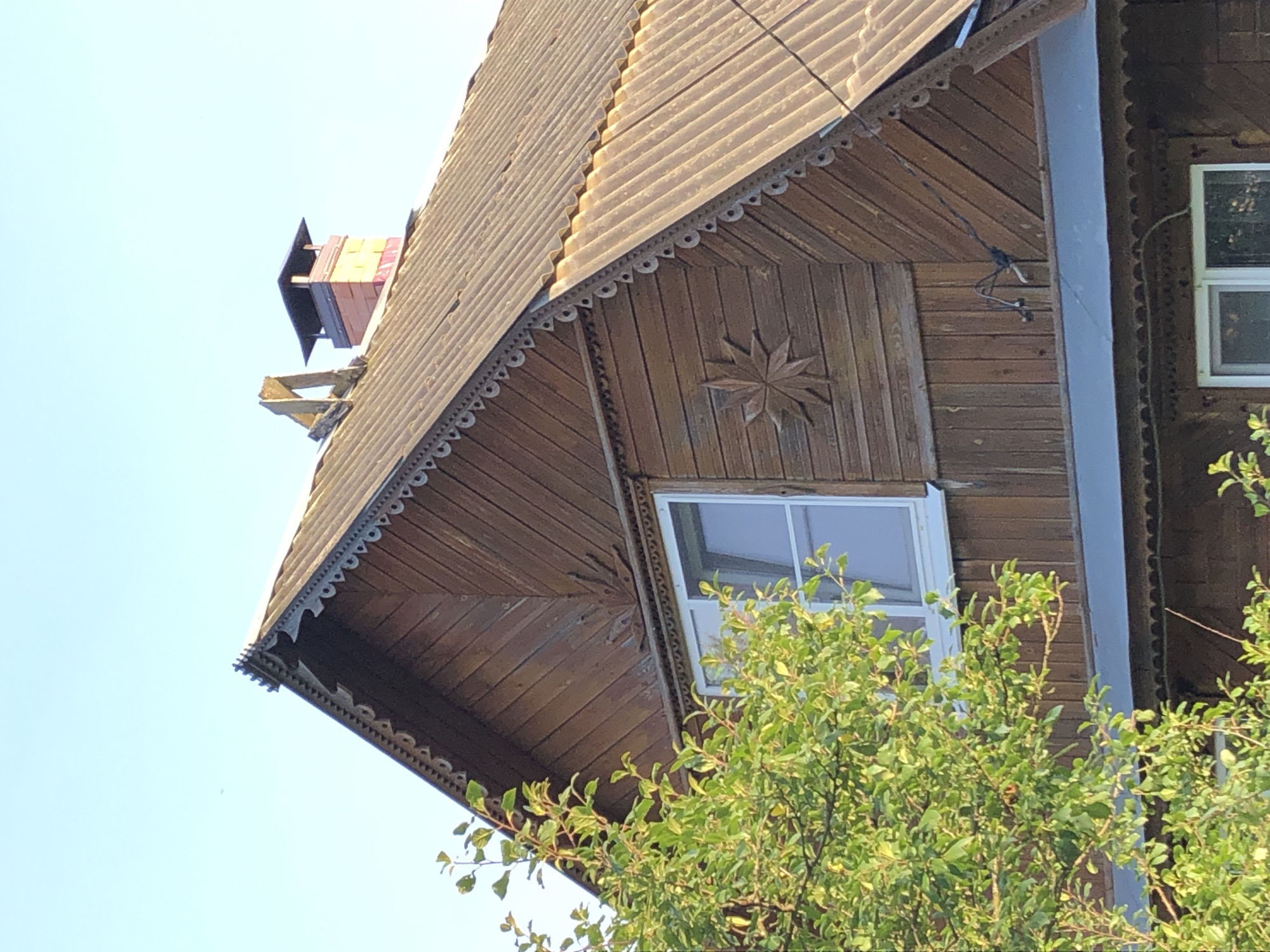 The image shows a close-up view of the upper part of a wooden house in Makeikha, Russia. The house features a gabled roof covered with corrugated material and adorned with decorative wooden trim along the eaves. There is a window on the upper level, and another window is partially visible on the lower level. A chimney made of brick with a dark cap rises from the roof. A tree with green leaves partially obscures the left side of the house. The sky is clear and blue.