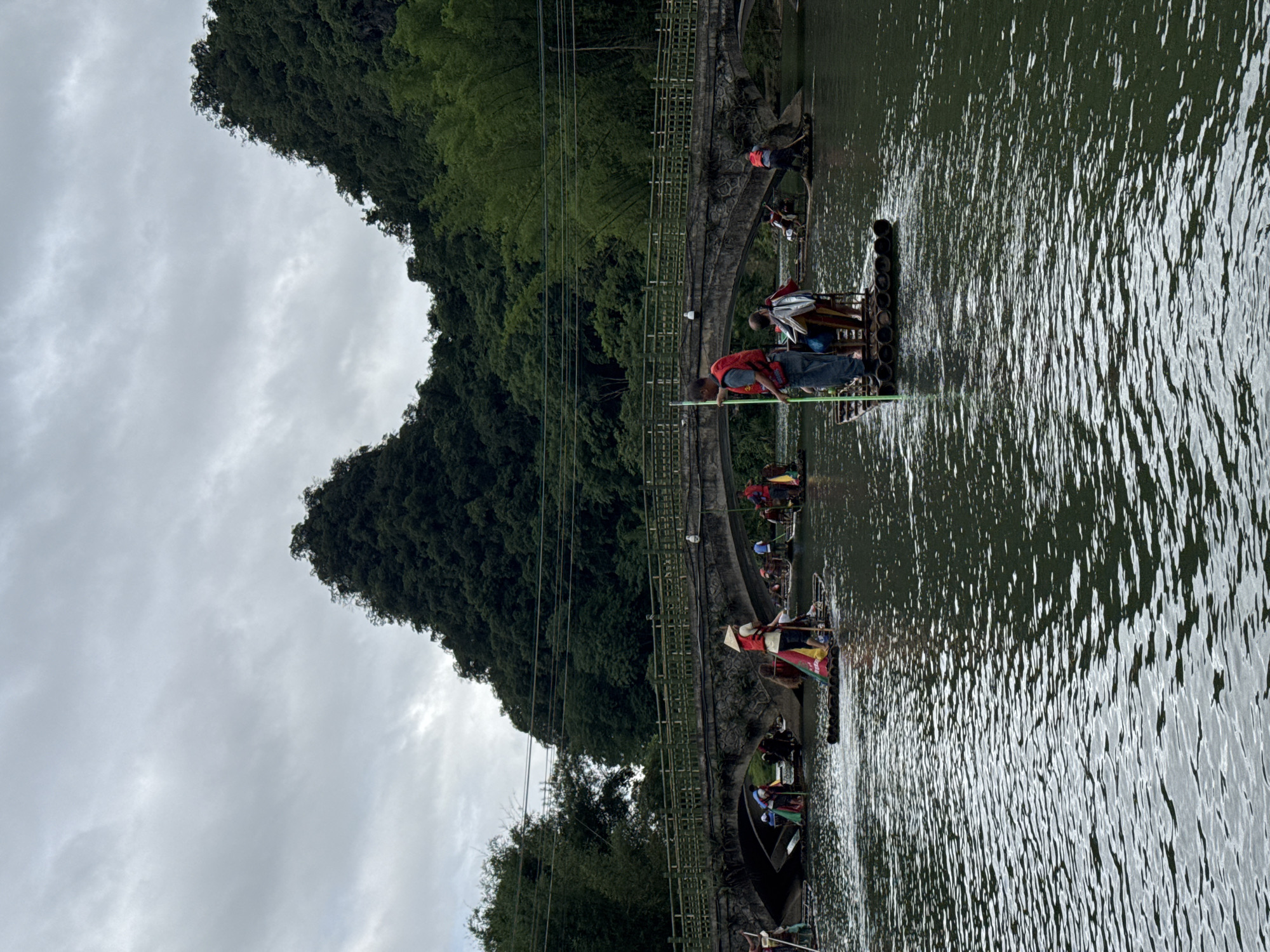 The image depicts rafts on the Yulong River in Guilin, China. Several rafts are visible on the river, each carrying people. The rafts appear to be propelled by individuals using poles to push against the riverbed. Lush green hills rise from the riverbanks, contrasting with the overcast sky. A bamboo fence runs along the edge of the riverbank, partially obstructing the view of the river.