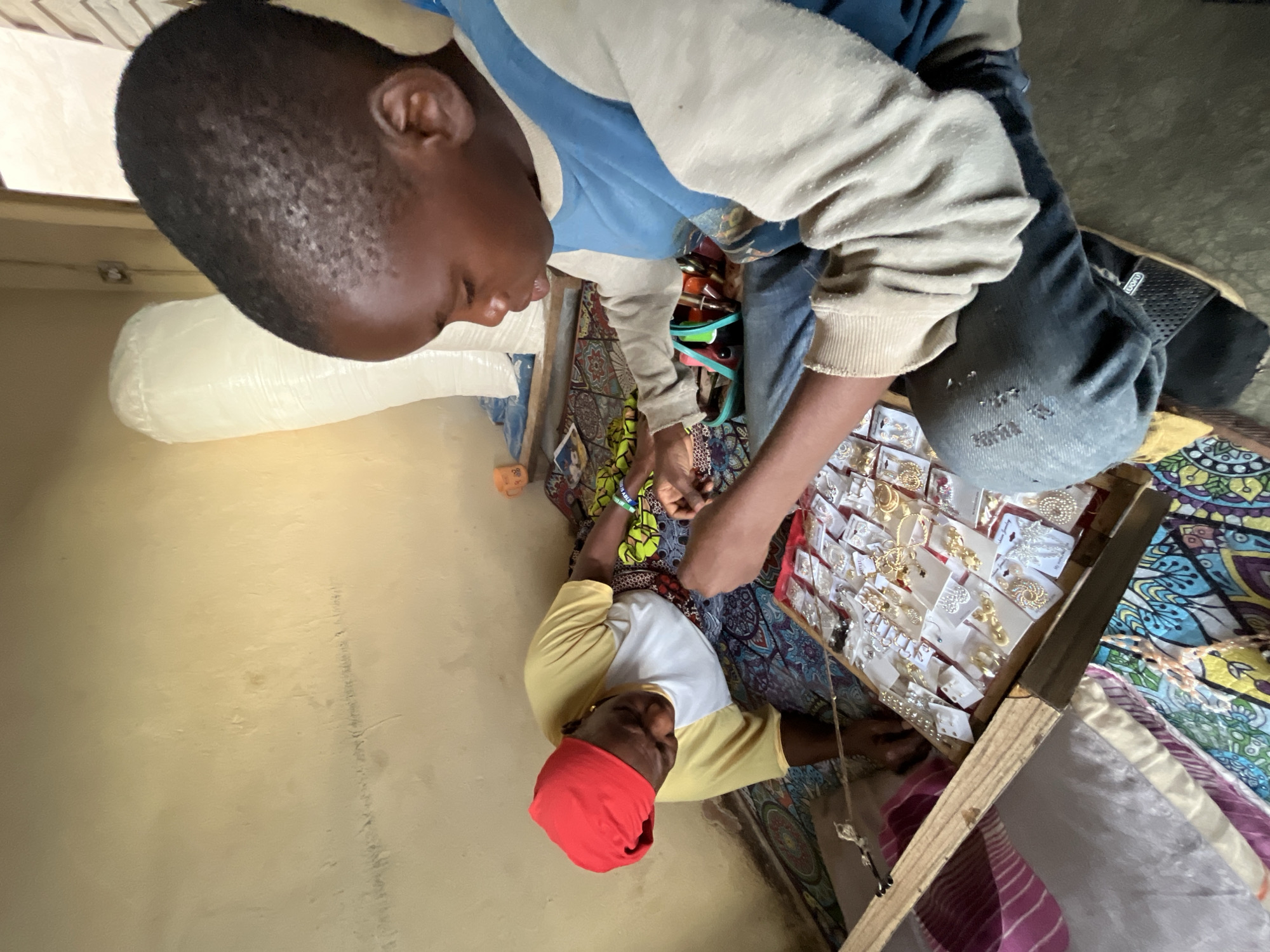 A scene in Jos, Nigeria, shows two individuals examining jewelry displayed on a patterned cloth. A child in a blue and tan sweatshirt is focused on items held in his hand, while an adult wearing a red head covering looks at a jewelry display. The display is on a slanted wooden stand. The room has a light yellow wall.