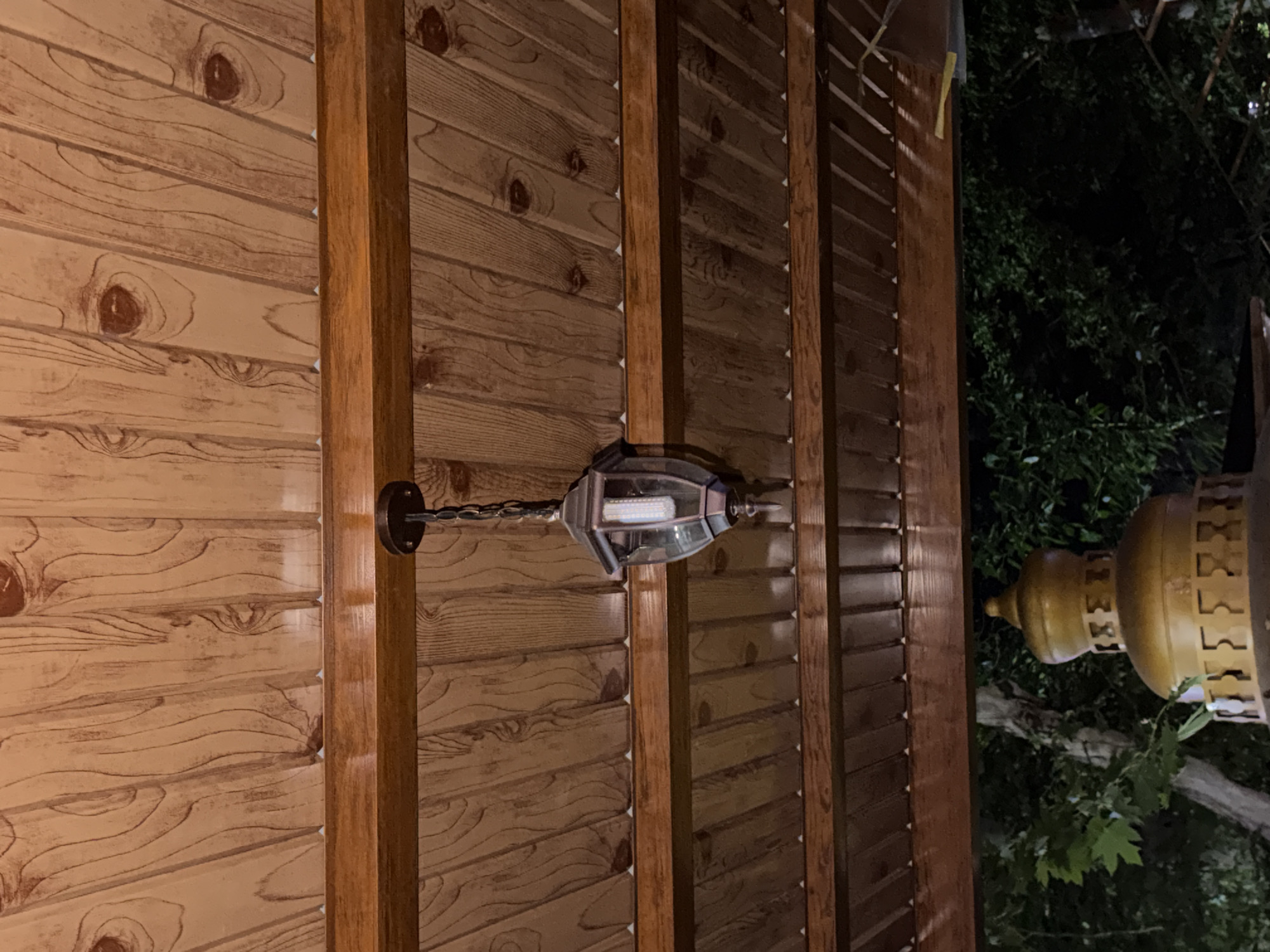 This is a medium-angle, eye-level shot of a wooden pergola in Chirchik, Uzbekistan, at night. The pergola features a wood-paneled ceiling with visible grain patterns, supported by wooden beams. A lantern hangs from the ceiling by a chain. Below the pergola, there is a golden, dome-shaped structure, possibly part of a larger decorative element or fountain. Lush green foliage and trees are visible in the background, suggesting an outdoor setting. The lighting is dim, characteristic of nighttime. No people are present in the image.