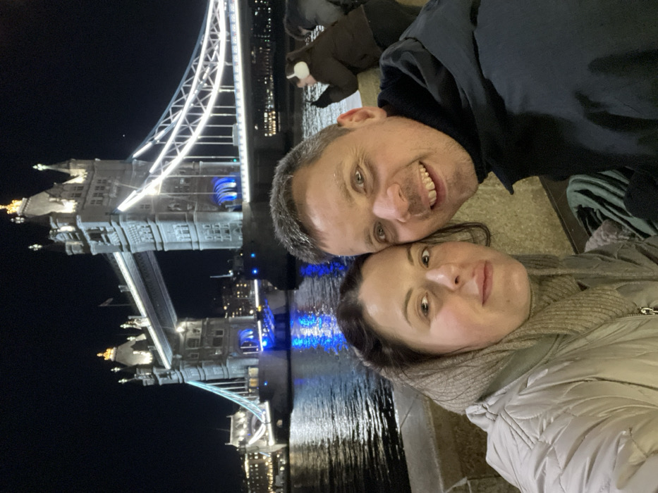 A selfie captures a man and a woman against the backdrop of London's Tower Bridge at night. The bridge's architectural details and illuminated structures are clearly visible in the background. The man smiles, while the woman has a neutral expression. The dark sky contrasts with the bridge's bright lights and reflections on the water.
