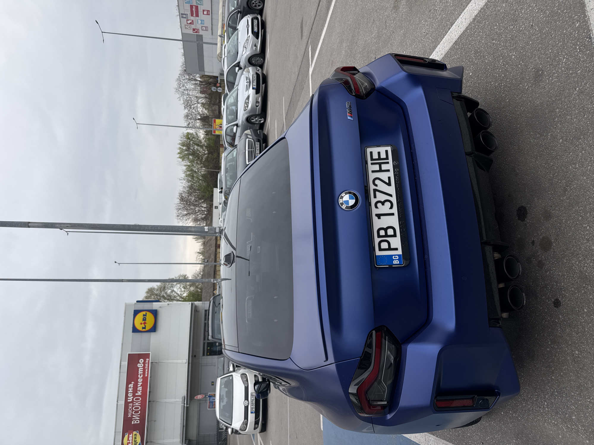 The image shows the rear of a matte blue BMW M2 parked in a parking lot. The license plate reads "PB 1372 HE" and indicates it's registered in Bulgaria. In the background is a Lidl store with a sign that reads "ниска цена, ВИСОКО качество". A white Volkswagen Golf is parked nearby, bearing the license plate "PB 8896 HP." Several other cars are parked in the distance. The location is presumably near a Lidl store in Plovdiv, Bulgaria, based on the language used on the signs and license plate details. The sky is overcast.