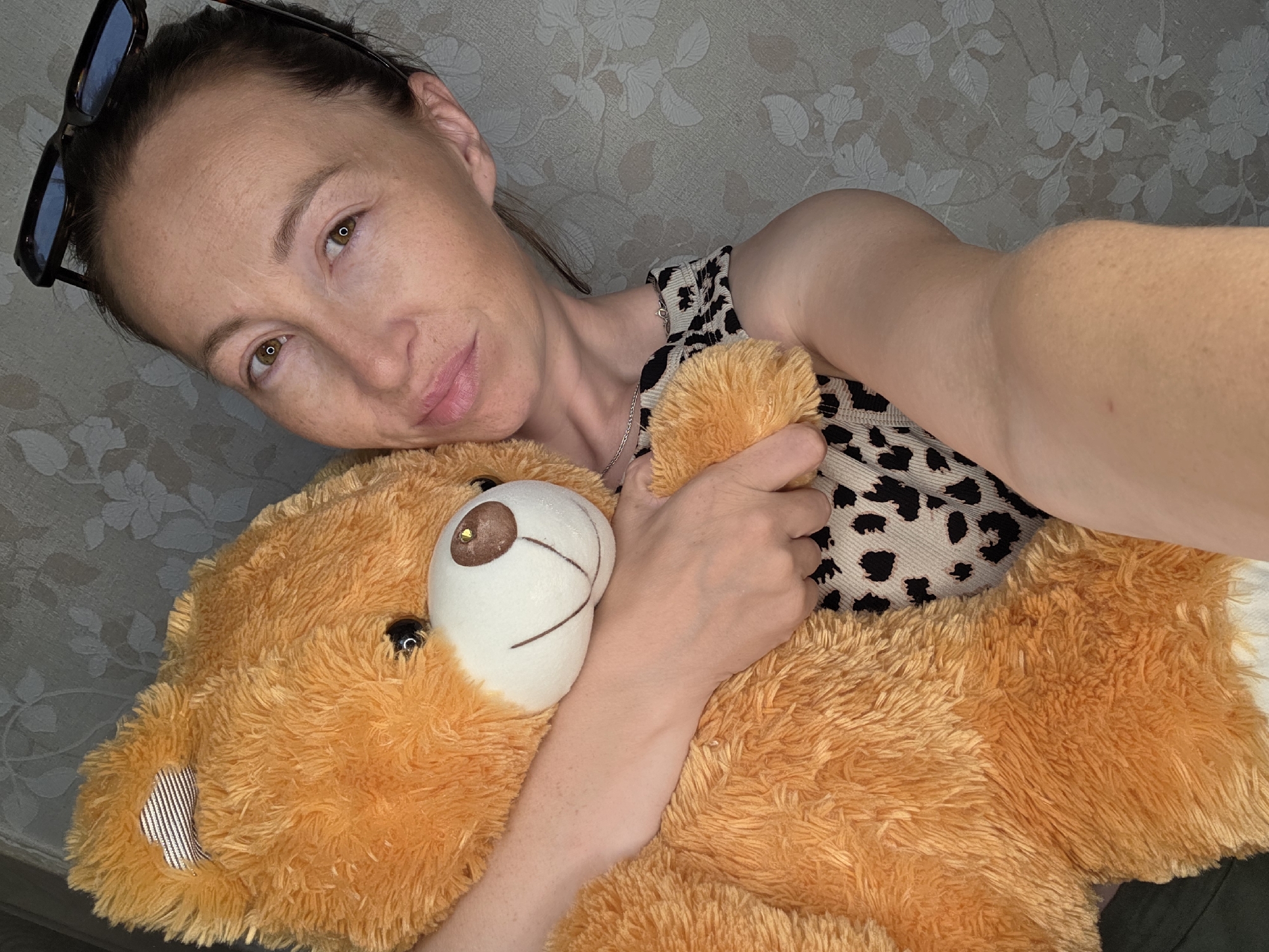 A woman with sunglasses perched on her head is seen from a high angle, lying down and holding a large, orange teddy bear. She is resting her head on the bear, with her left arm wrapped around it. The woman has brown hair and brown eyes, and she is looking towards the camera with a slight smile. She is wearing a leopard-print top. The teddy bear is fluffy and has a white face with brown eyes and a stitched nose. The background appears to be a wall with a floral wallpaper pattern. The lighting suggests it could be daytime indoors. The overall mood of the image is relaxed and affectionate. There is no visible text in the image.