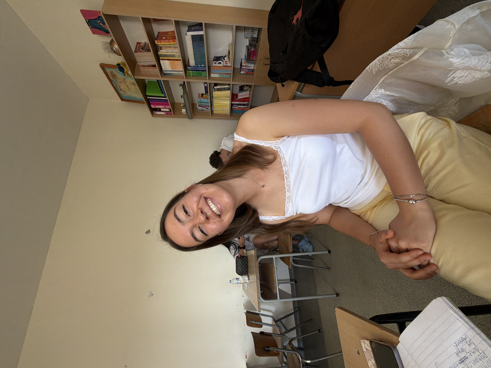 The image depicts a young woman with long brown hair, wearing a white tank top and yellow pants, sitting at a desk in a classroom. She is smiling directly at the camera, with her hands clasped in her lap. The classroom appears to be old, with wooden desks and chairs, and a bookshelf filled with books in the background. There are other students in the background, also seated at desks. The overall lighting suggests it is daytime, likely indoors. The specific location in Burgas, Bulgaria cannot be determined from the image alone.