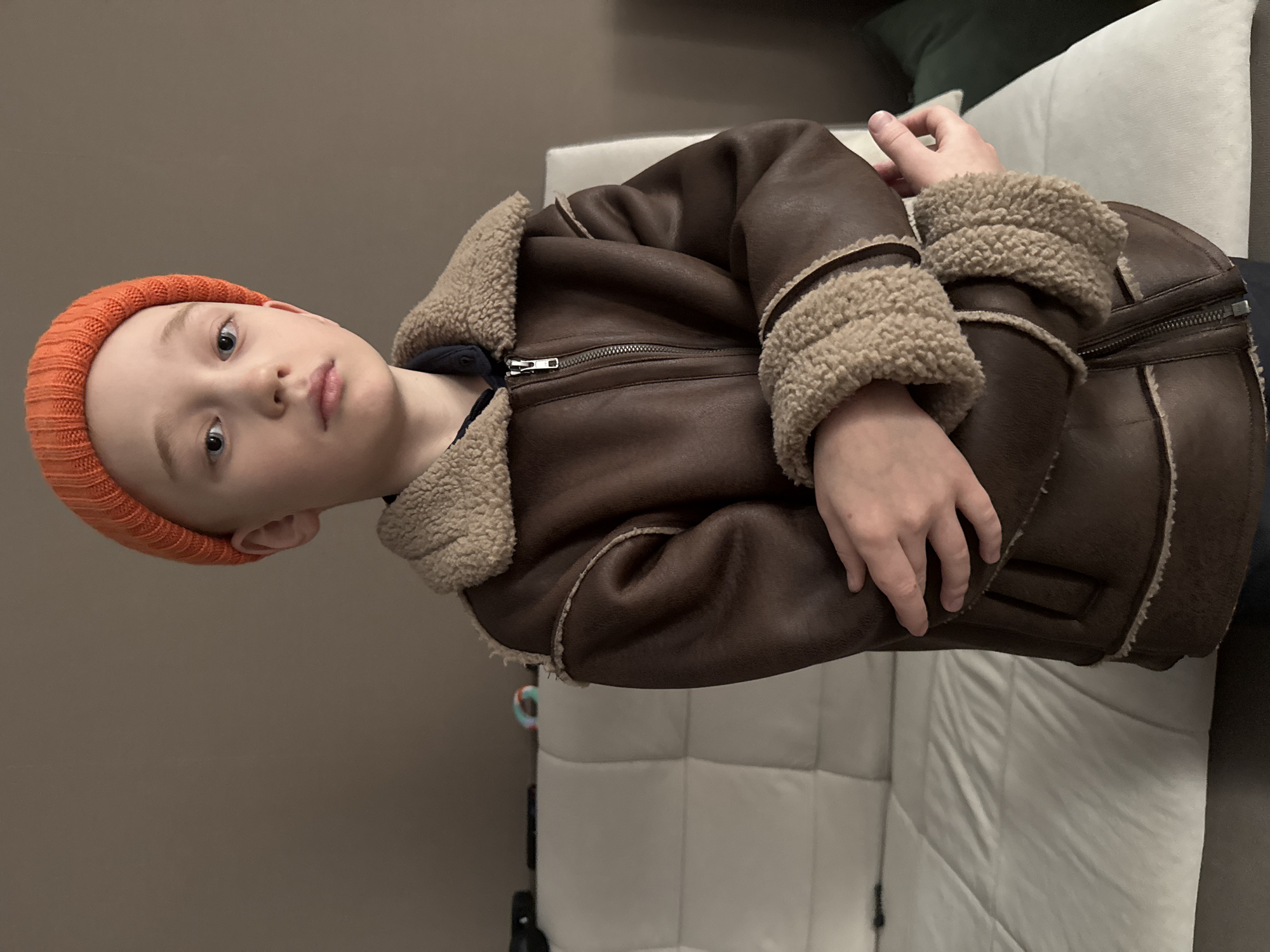 A young boy, estimated to be between 6 and 10 years old, is depicted in an indoor setting within Votkinsk, Russia. He stands facing the viewer with a neutral facial expression and direct gaze from his blue eyes. The boy wears an orange knitted beanie and a brown shearling-lined jacket, which features a prominent metal zipper and light beige fleece trim on the collar, cuffs, and seams. A dark blue collar from an underlying shirt is visible at the neckline. His arms are crossed at approximately waist height, with hands partially obscured by the jacket sleeves. The background features a plain light-brown wall dominating the upper and left portions of the frame, contrasting with a light-colored, multi-cushioned sofa or couch positioned to the left and lower right. A dark object and a colorful circular toy are faintly visible on the sofa's upper-left side. The scene is illuminated by ambient, even lighting.