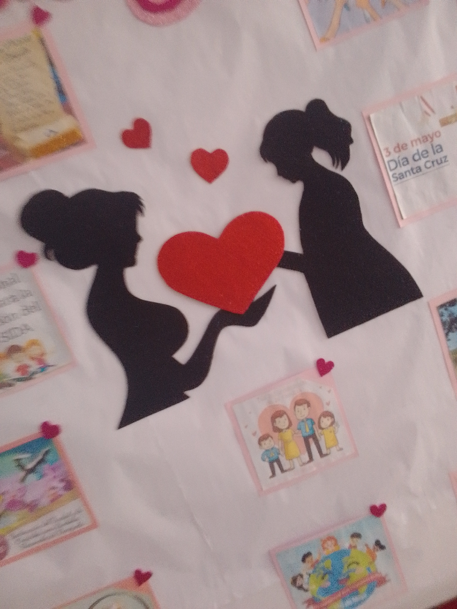 This media file depicts a bulletin board decorated with cutouts and small pictures, likely for a celebration or event. The central subjects are two black silhouettes of women, one larger than the other, embracing and holding a large red heart between them. Smaller red hearts are scattered around the silhouettes, along with a few small pink hearts attached to some of the pictures.
Scattered across the board are several small, square pictures. One picture shows a drawing of a family with two parents and two children, framed by a pink border and a pink heart. Another picture, also framed in pink, shows a colorful depiction of people surrounding a globe. A third picture, at the top right, has text in Spanish that reads "3 de mayo Día de la Santa Cruz". Another picture, at the top left, is partially obscured but appears to show text and an illustration. The background is a plain white surface, suggesting the bulletin board is indoors or in a covered area.
The overall theme appears to be love, family, and possibly a celebration related to May 3rd. The presence of hearts and family imagery suggests a theme of affection and togetherness. The silhouettes and bold heart cutout create a strong visual focus on the relationship between the two figures. The setting appears to be a communal space where such a display would be appropriate, such as a school or community center.