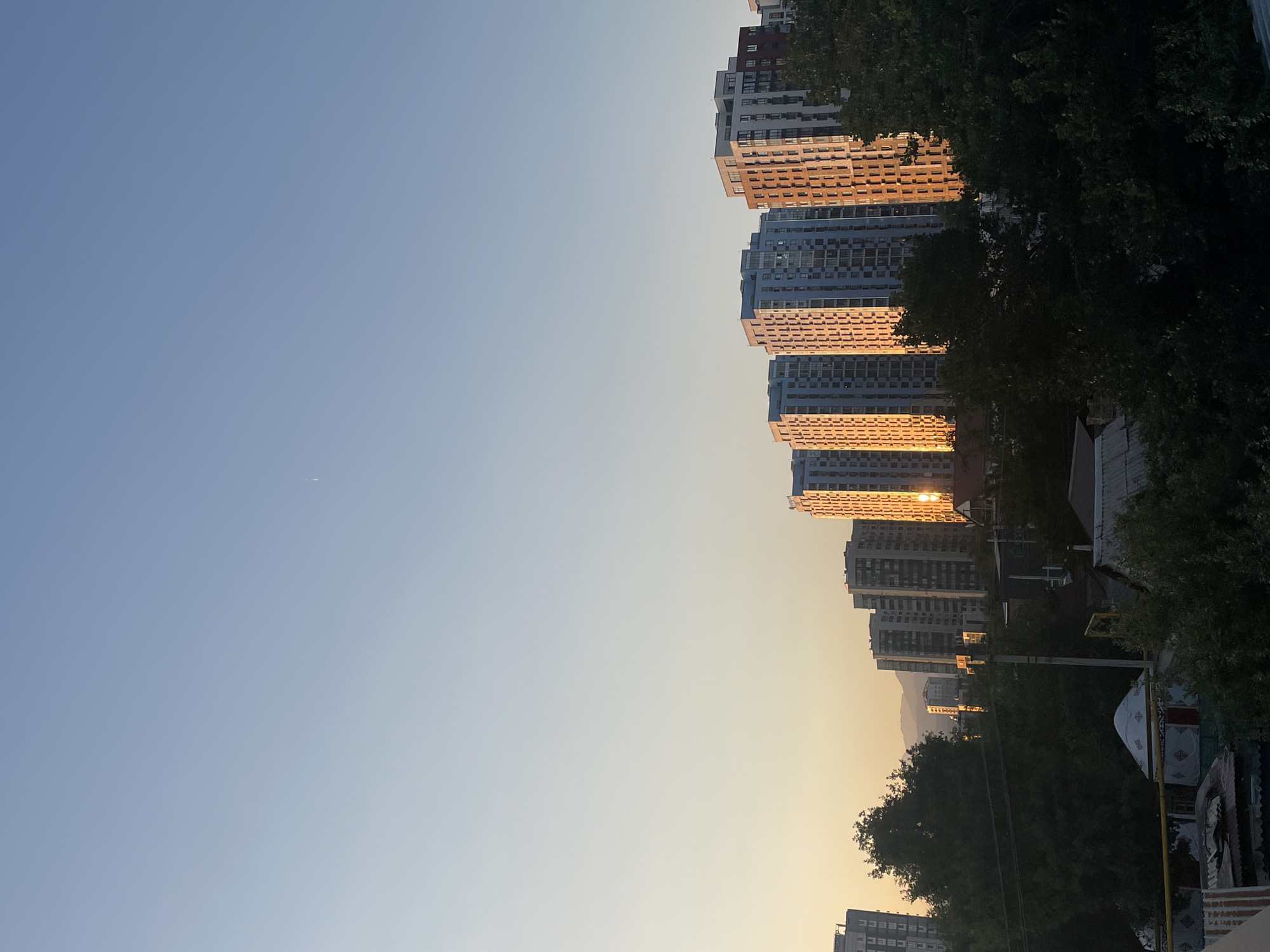 The image depicts a cityscape at sunset, likely in Almaty, Kazakhstan, given the presence of mountainous terrain in the background and the architectural style of the buildings. The foreground is dominated by lush green trees and lower-lying structures, including what appear to be yurts, indicative of residential areas. Several tall, modern apartment buildings are situated in the midground and background, with the setting sun casting a warm, golden light on their facades, creating a striking contrast with the clear, pale blue sky. A faint airplane contrail is visible in the upper portion of the sky. The overall scene conveys a sense of urban development blending with natural elements under the soft glow of sunset.