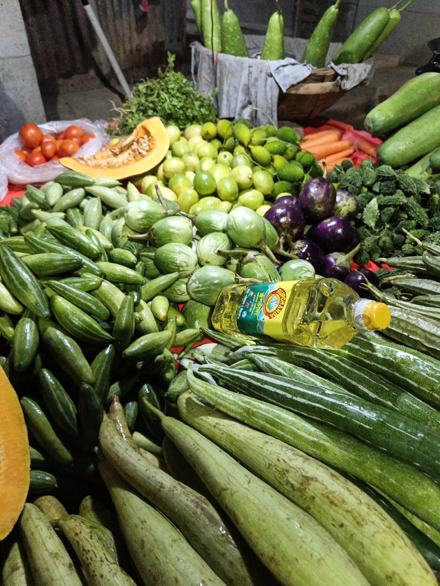 This image depicts a vibrant market stall in Dhaka, Bangladesh, overflowing with fresh produce. The scene is bustling with an array of vegetables arranged on a red surface. Prominently featured are long, green gourds, some with distinctive white stripes, alongside smaller, finger-like green vegetables that appear to be pointed gourds or ivy gourds. There are also round, light green vegetables, possibly small eggplants or some variety of gourd, and a few dark purple eggplants. A sliced piece of pumpkin, displaying its orange flesh and seeds, adds a pop of color. Interspersed among the vegetables are bright red tomatoes in a plastic bag and some carrots. A bottle of cooking oil with a yellow label and cap rests on top of some of the vegetables. In the background, more long green gourds are visible, some standing upright in a basket lined with cloth. The overall impression is one of abundance and the lively atmosphere of a local marketplace. The lighting suggests it could be daytime, possibly late afternoon given the warm tones. There are no people visible in the frame, but the sheer volume and variety of produce imply a seller-buyer interaction is imminent or ongoing. The label on the oil bottle has some visible text in a language that appears to be Bengali.