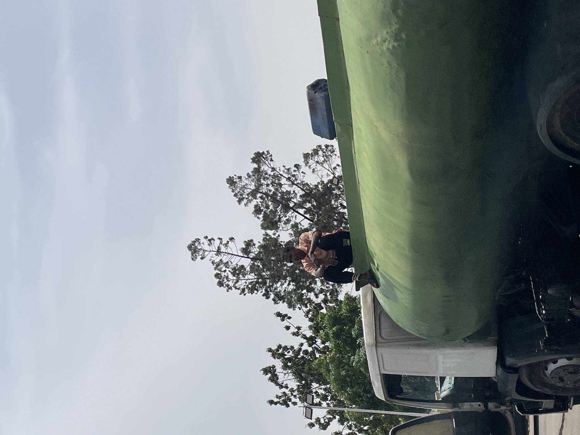 A man sits atop a large, green tanker truck. The truck is parked near trees. The sky is overcast. The location is Jos, Nigeria.