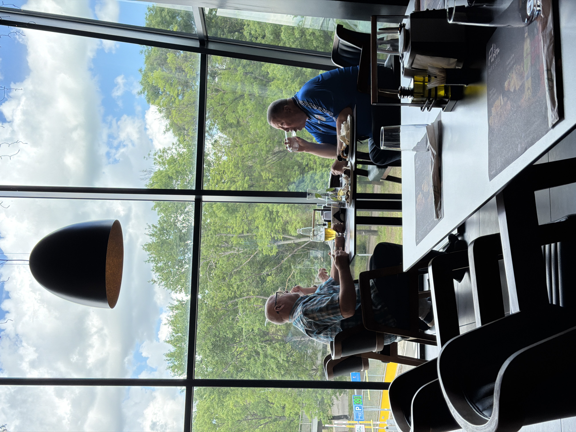 The image shows two men dining at a table inside a restaurant with large windows overlooking an outdoor view. One man wears a blue shirt and is seen drinking from a glass. The other man wears a checkered shirt and is also eating. On the table are plates of food, drinks, and restaurant menus. Outside the window, green trees and a cloudy blue sky are visible. There's also a parking sign with the Cyrillic letters "Р" and the number "401". The image appears to have been taken at the Flora Bar & Grill in Burgas, Bulgaria.