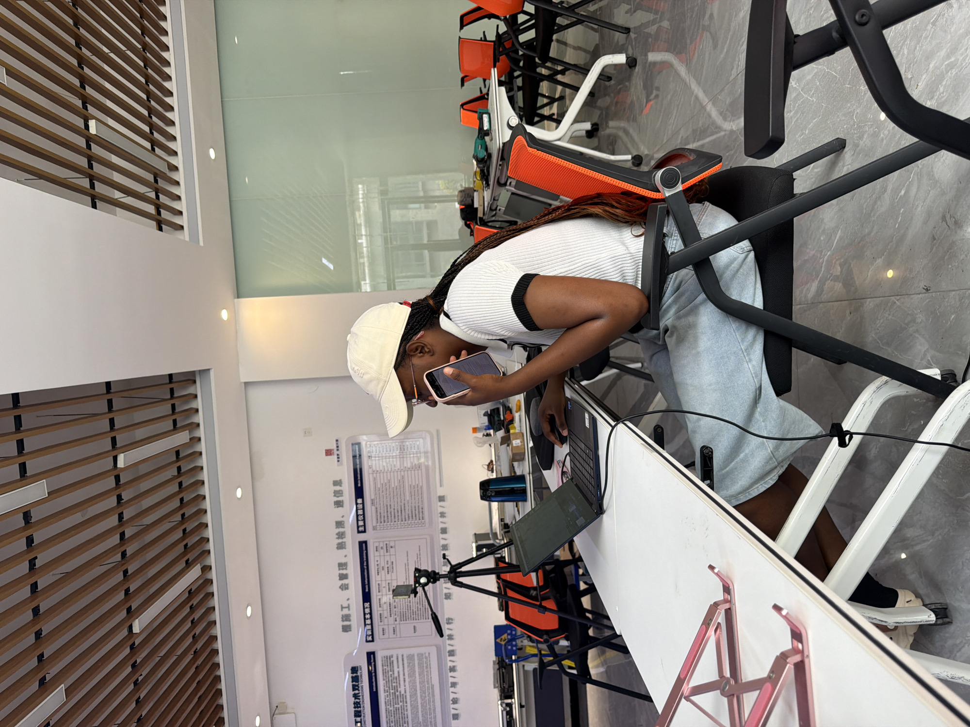 A young Black woman wearing a white baseball cap, glasses, and a white short-sleeved shirt with black trim is seated at a white desk, talking on her smartphone. She is in a modern office or training room with a grey tiled floor and glass partitions in the background. The room contains several tables with orange-accented chairs. A laptop is open on the desk in front of her, and a tripod with a camera is also visible. The woman's hair is braided and extends down her back. The desk has a pink laptop stand on it. The location is identified as Jiangjin, China.